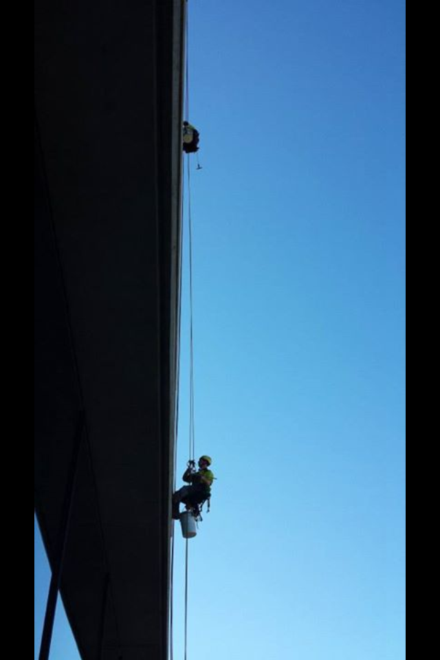 A worker wearing safety gear is rappelling down the side of a building, held by ropes with a bucket attached to their harness, against a clear blue sky.
