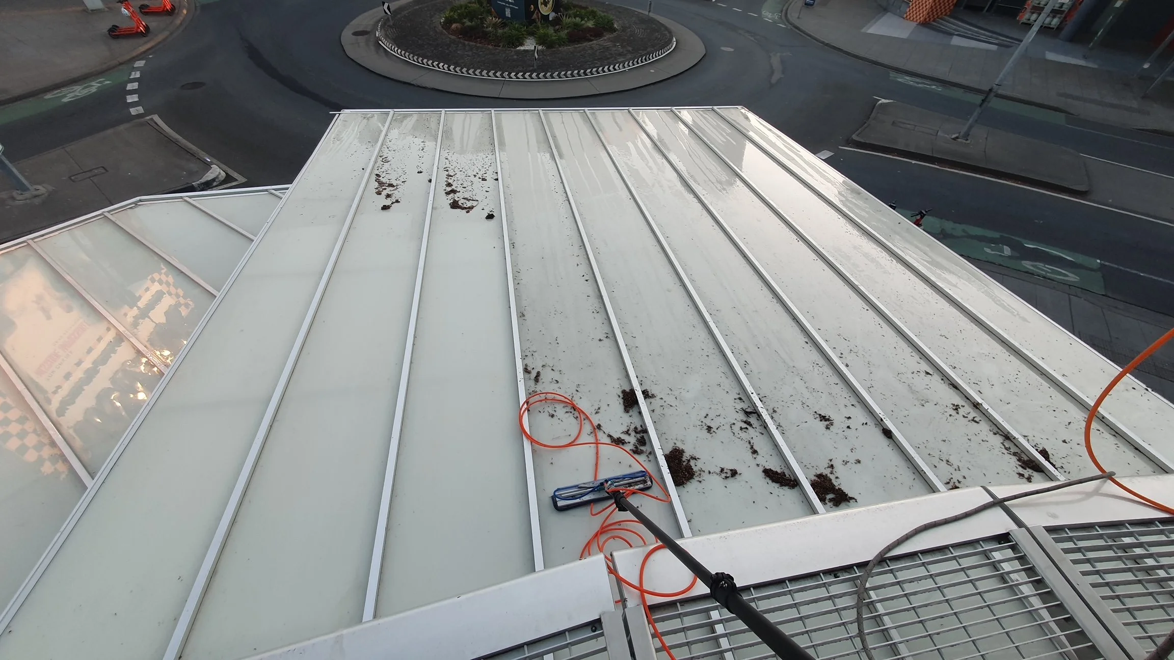 Dirty white metal roof with debris and dirt, seen from an elevated angle, with cleaning equipment and a long orange extension cord.