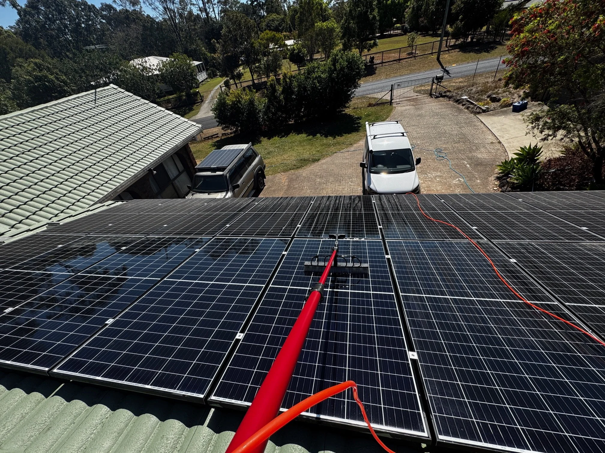 Looking down at solar panels installed on a roof with a camera or robotic cleaner in the center, and multiple cars parked in a driveway below, with trees and a street in the background.