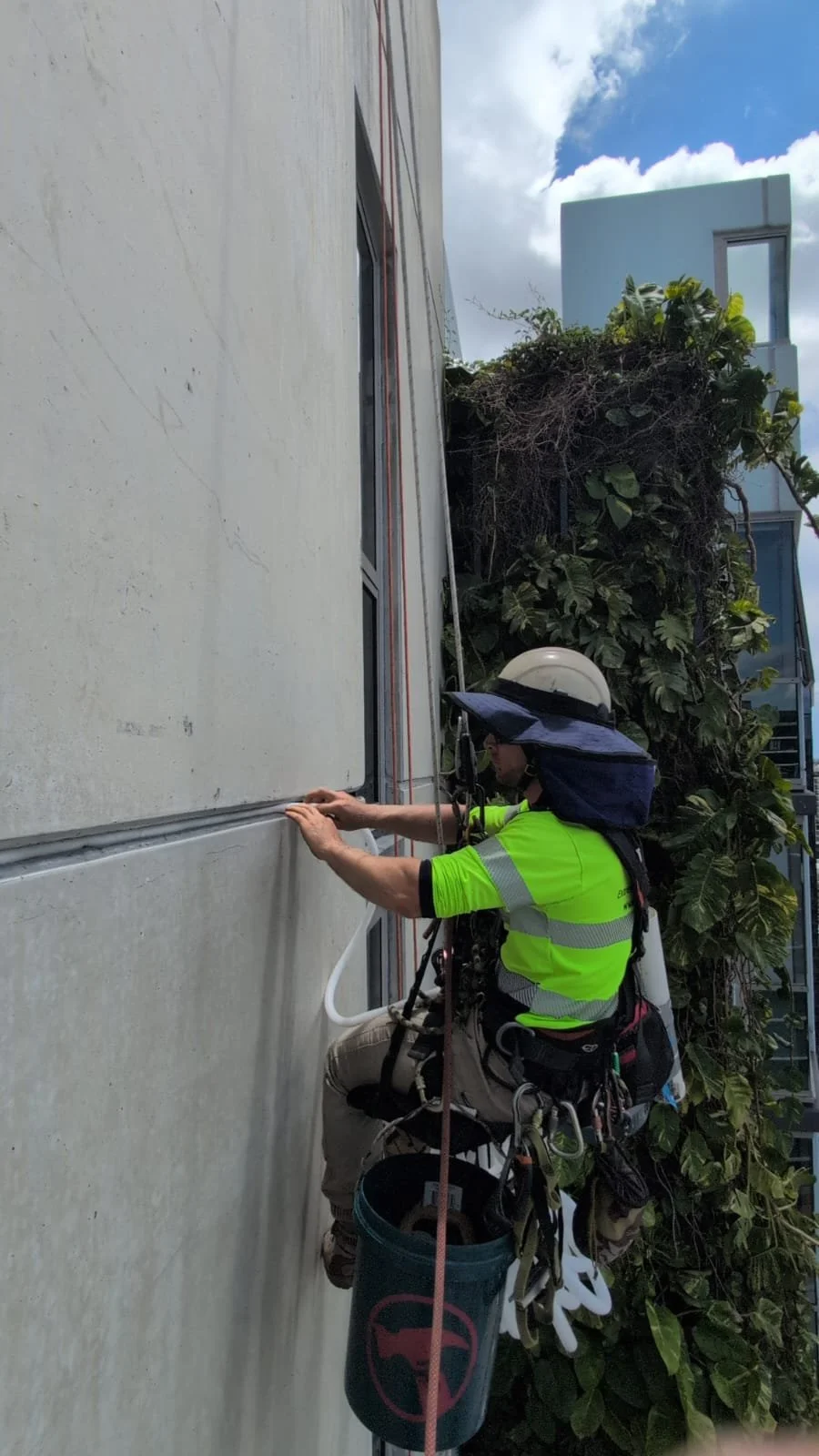A worker wearing a helmet, sunglasses, and a neon yellow safety shirt is installing or inspecting electrical wiring on the side of a building, with greenery and a blue sky with clouds in the background.