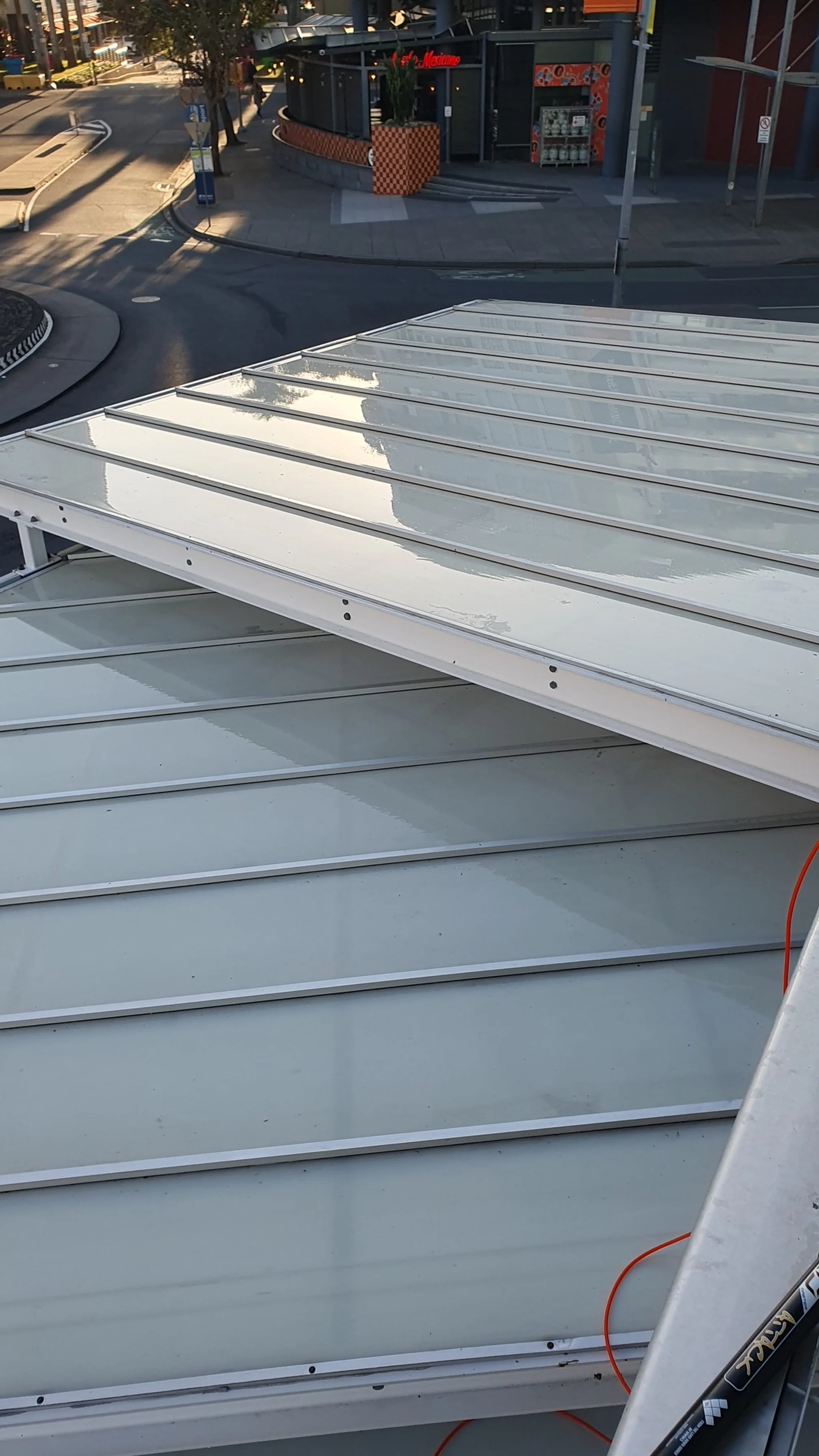 Close-up of a solar panel rooftop with a city street in the background, showing a building with a red sign, trees, and street signs.
