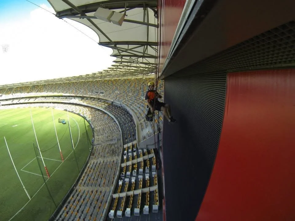 Worker hanging on the exterior wall of a stadium during construction or maintenance, with a view of the playing field below.