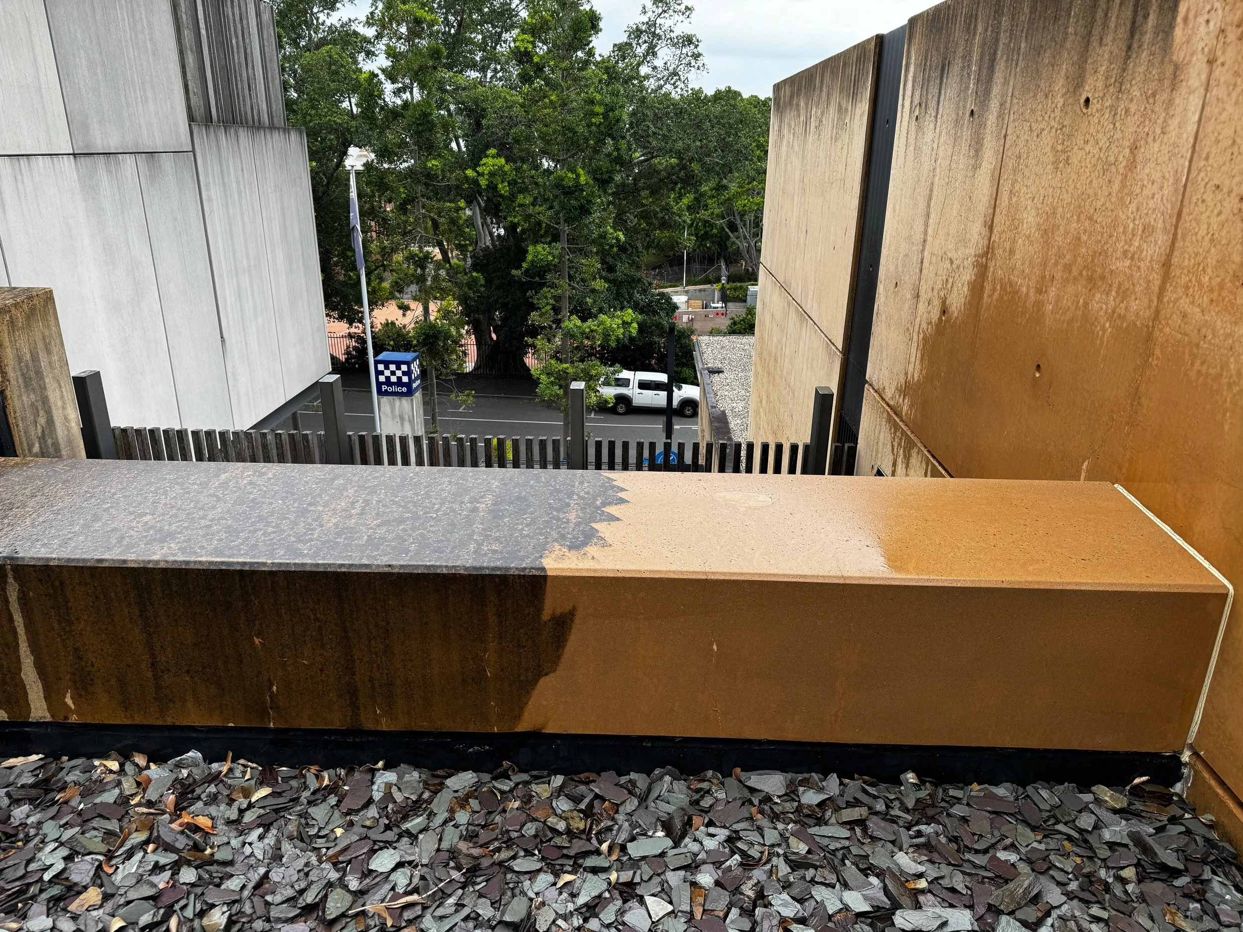 Overview of a rooftop with two rust-colored metal panels, a gravel surface, and a city street in the background featuring a Police station sign, trees, and parked cars.