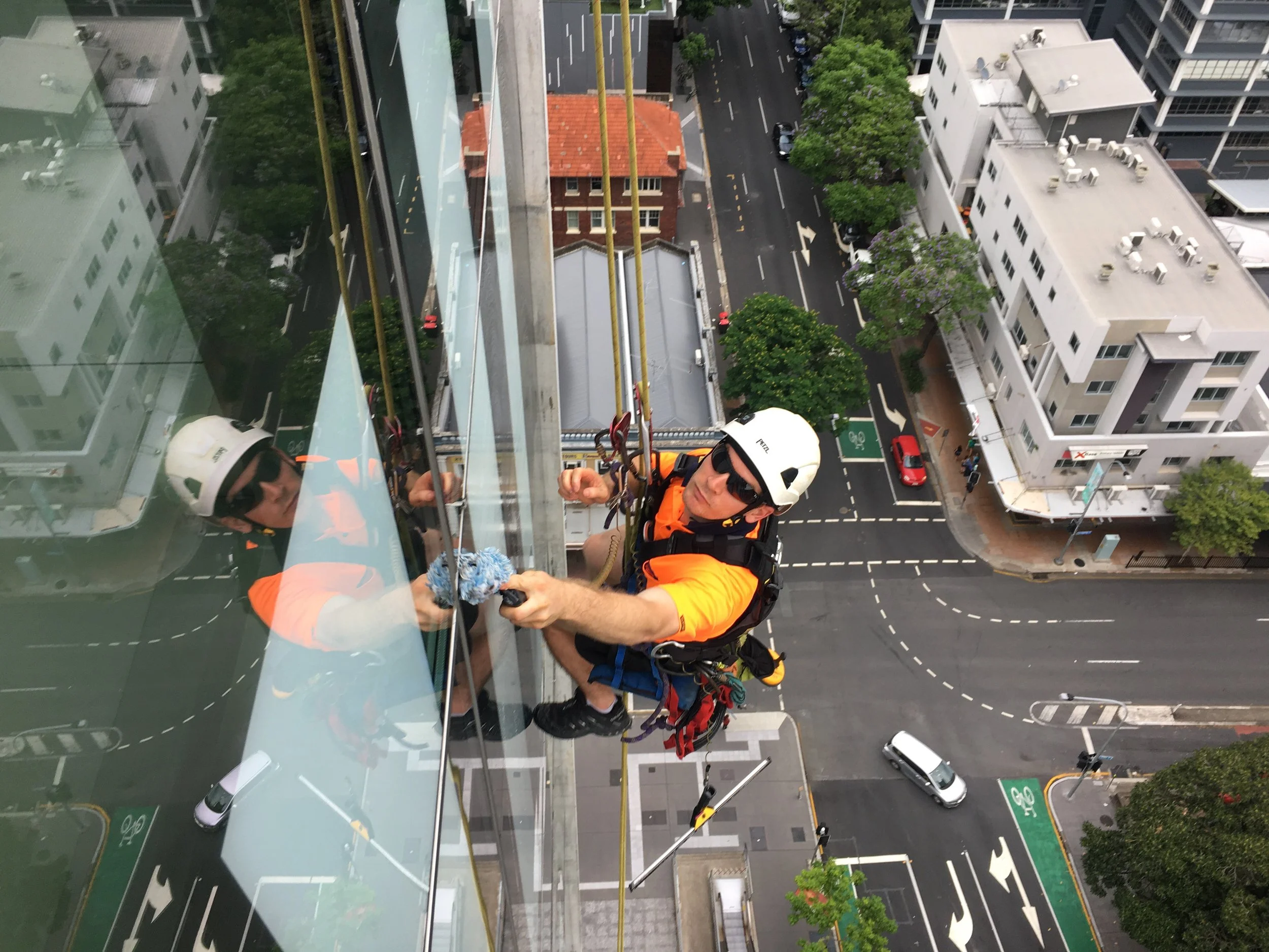 Two workers wearing helmets and safety gear cleaning the exterior glass of a tall building using a cloth and rope.