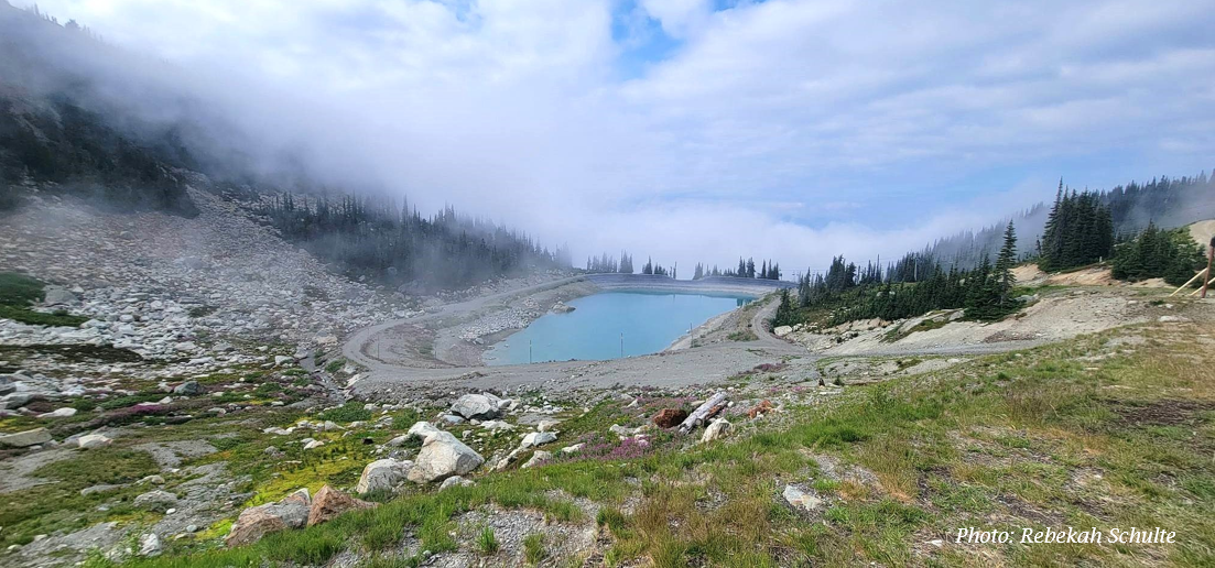 Mountain landscape with a mountain pool or lake, surrounded by rocky terrain, evergreen trees, and fog or low clouds in the background.