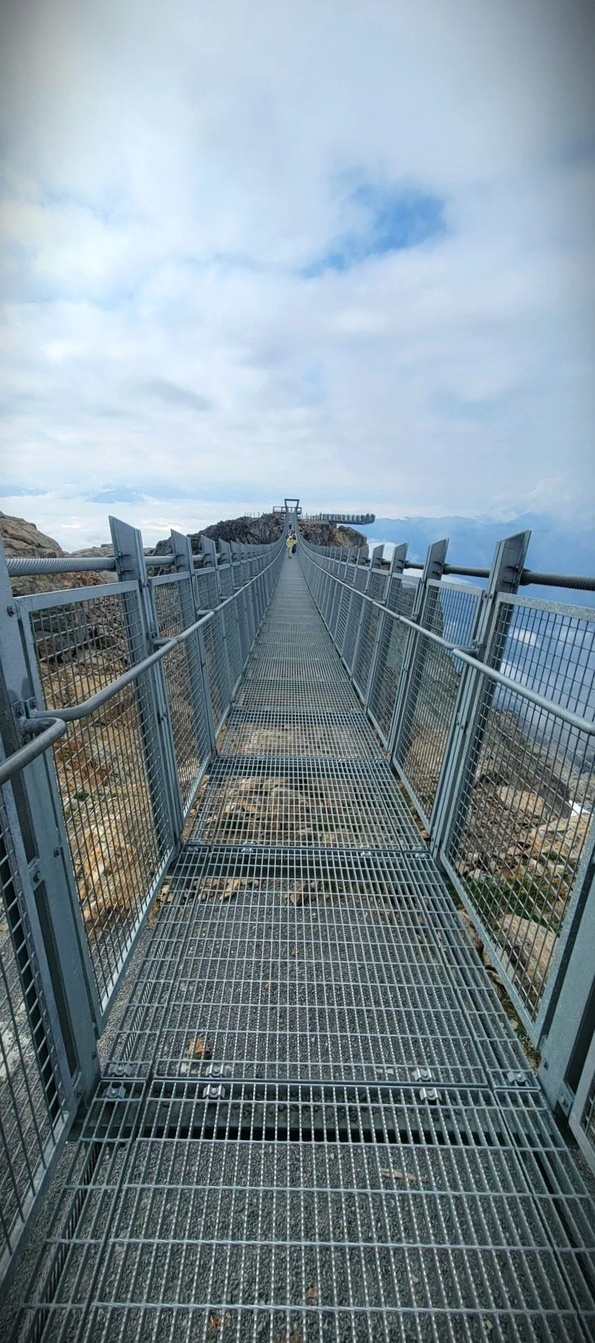 Metal walkway bridge with railings, extending towards a mountain summit under cloudy sky, with distant mountain range in the background.