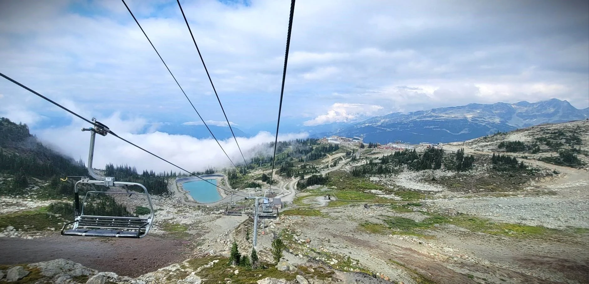 A mountain landscape viewed from a ski lift, with a cloudy sky, a lake, and distant mountain peaks.