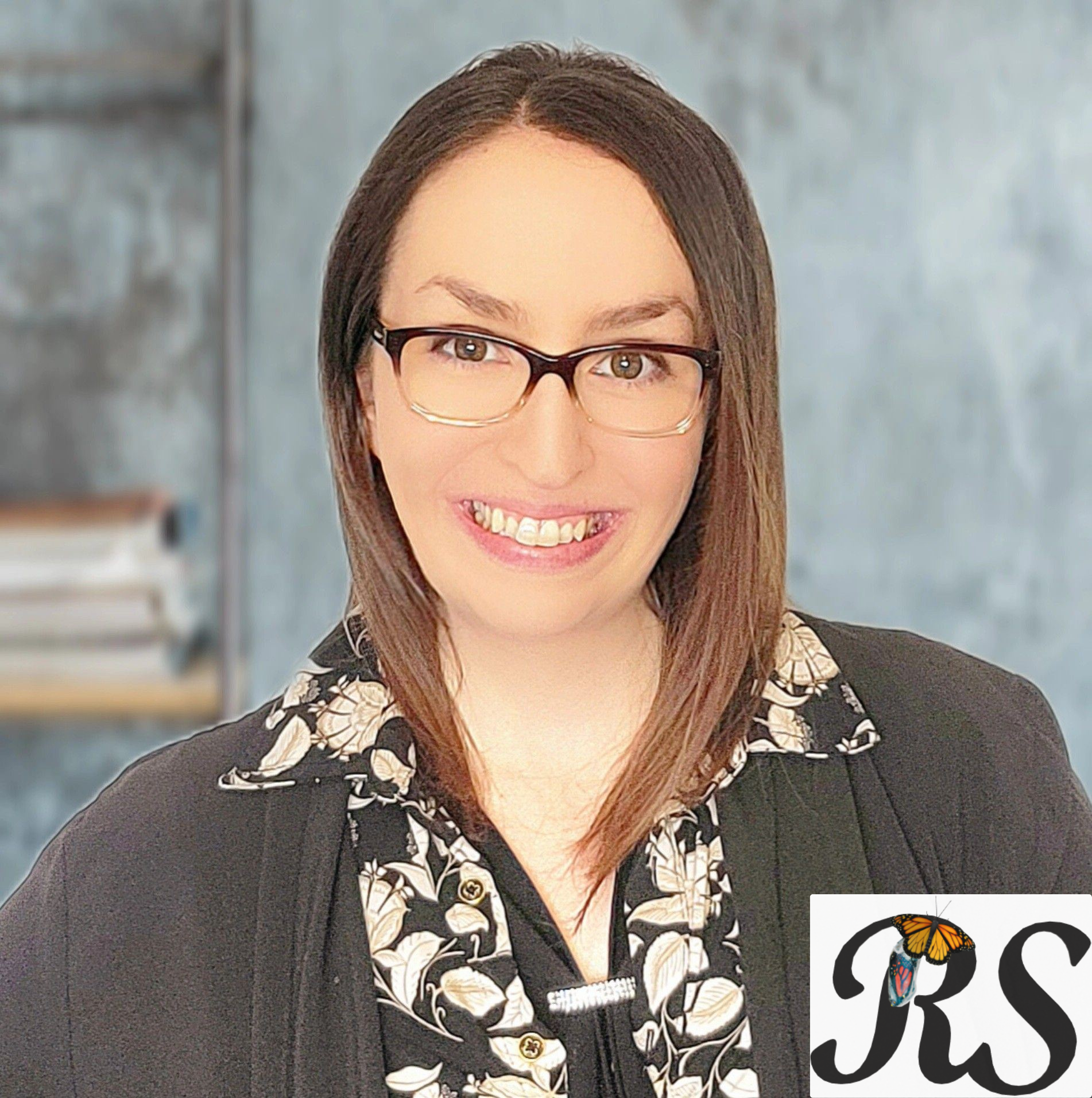A woman with shoulder-length brown hair, glasses, and a cheerful smile, wearing a black and white floral patterned top and black blazer, standing in an indoor setting with a blurred background