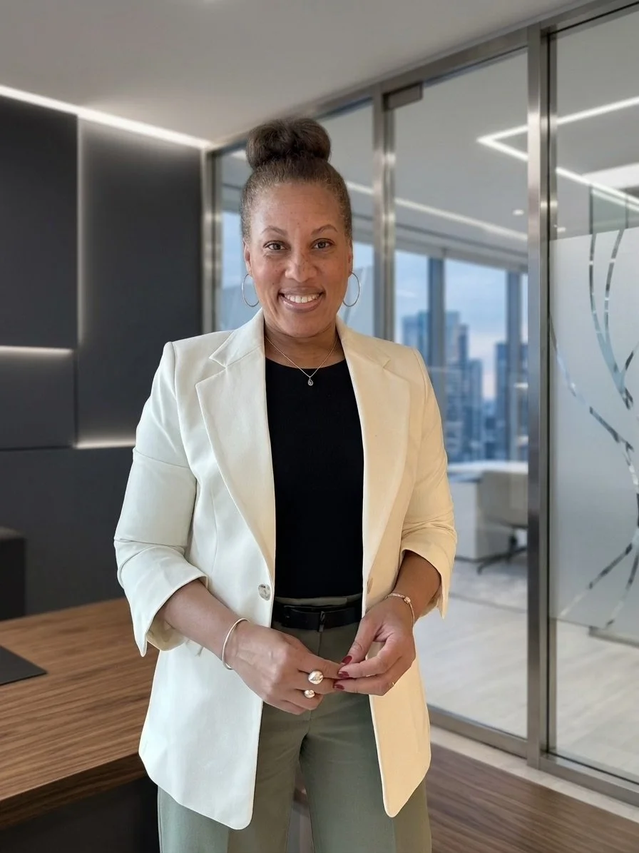 Smiling woman in a cream blazer and black top standing in a modern office with large windows and a city view.
