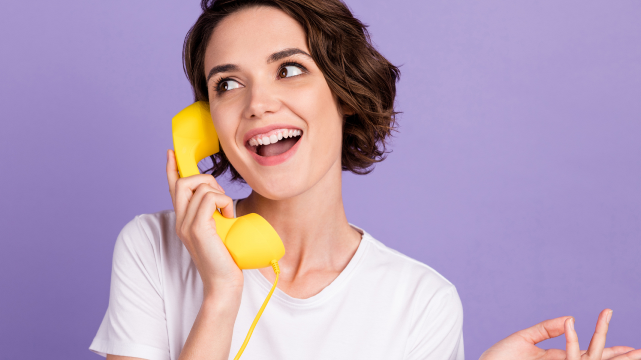 A young woman with short brown hair smiling while talking on a yellow telephone against a purple background.
