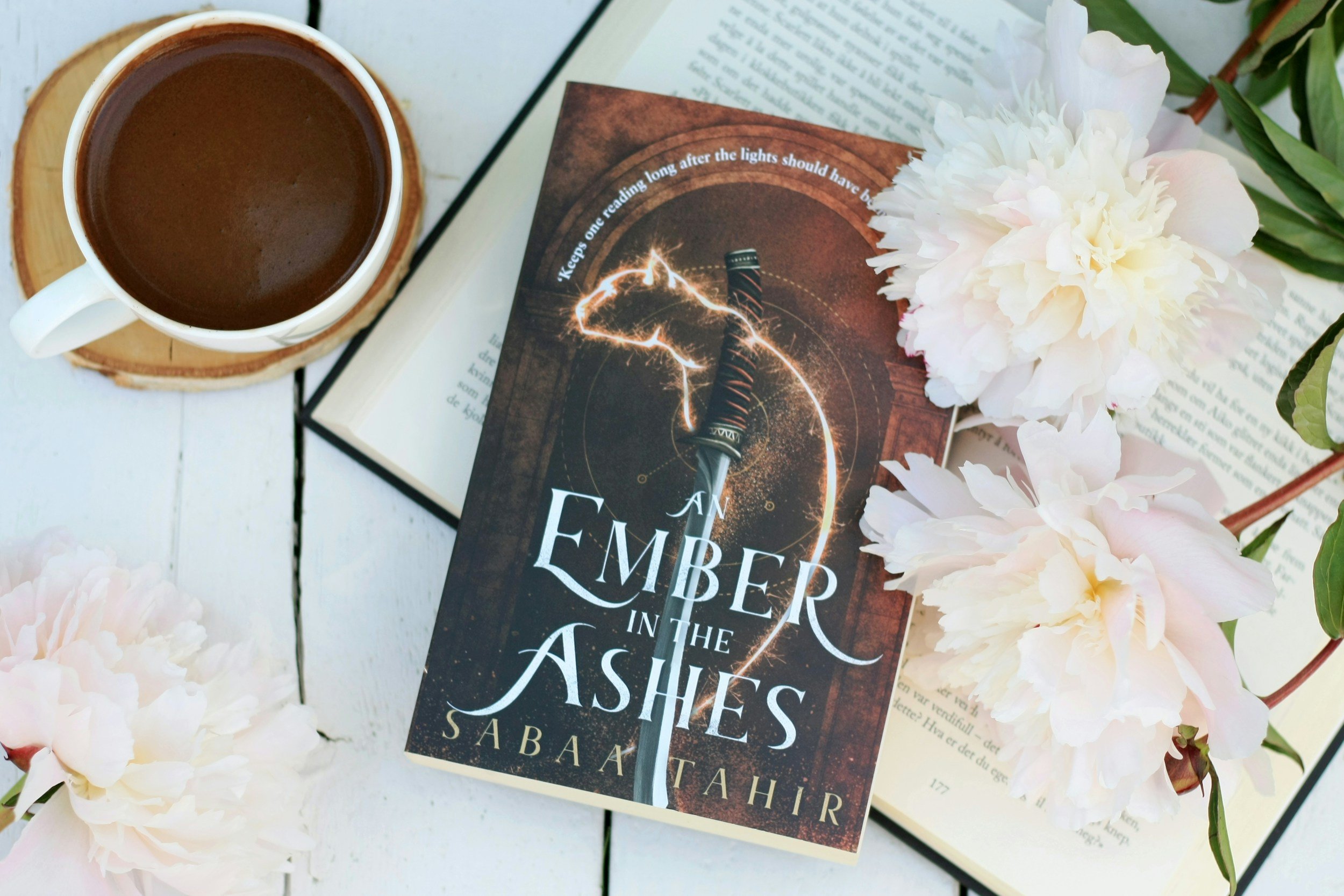 A white cup of hot chocolate on a wooden coaster, beside a partially open book and a copy of the novel 'An Ember in the Ashes' by Sabaa Tahir, surrounded by white and pink flowers on a white surface.