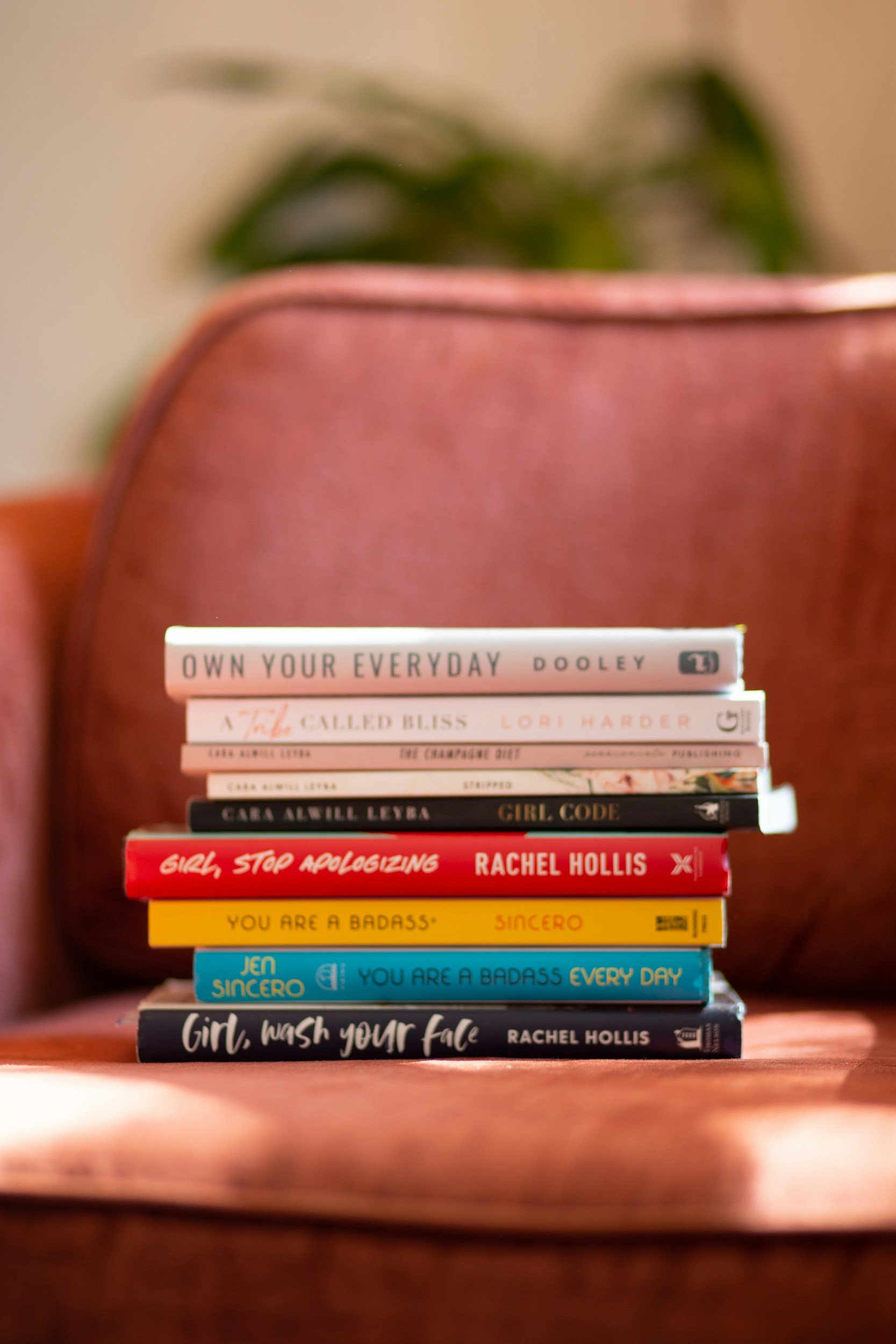 Stack of books on a pink armchair, with a blurred background of green plants. The books include titles such as 'Own Your Everyday,' 'Girl, Wash Your Face,' 'Girl Code,' and 'Savor.'