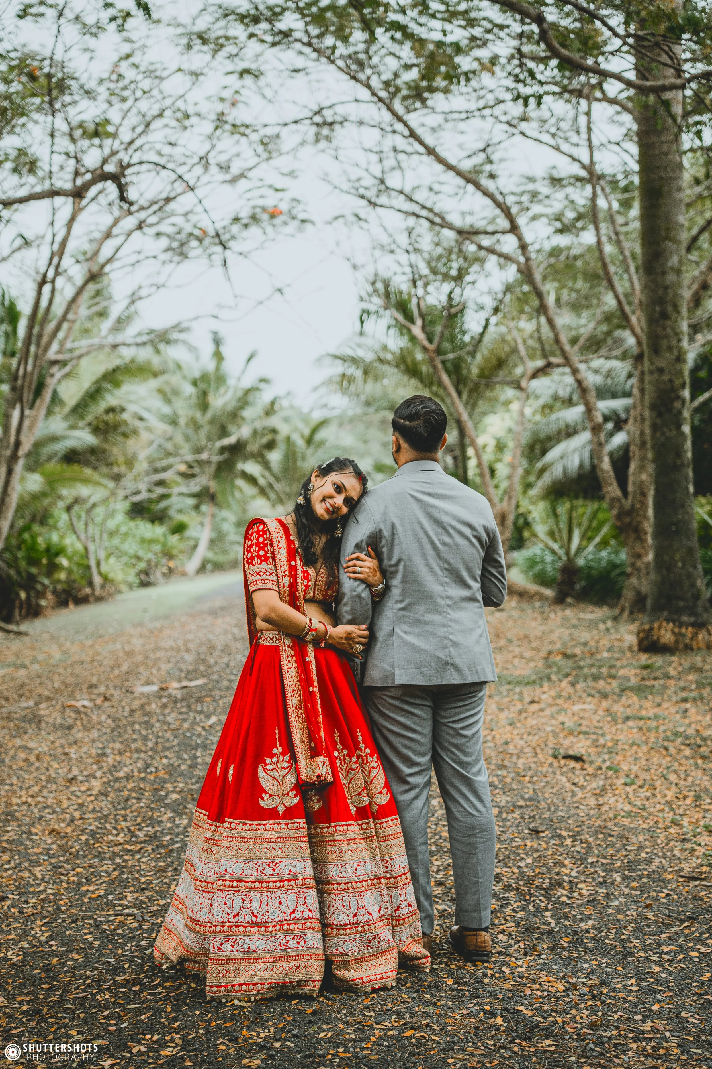 A couple standing on a dirt path surrounded by trees. The woman wears a red embroidered traditional dress and the man wears a gray suit. The woman smiles and leans her head on the man's shoulder.