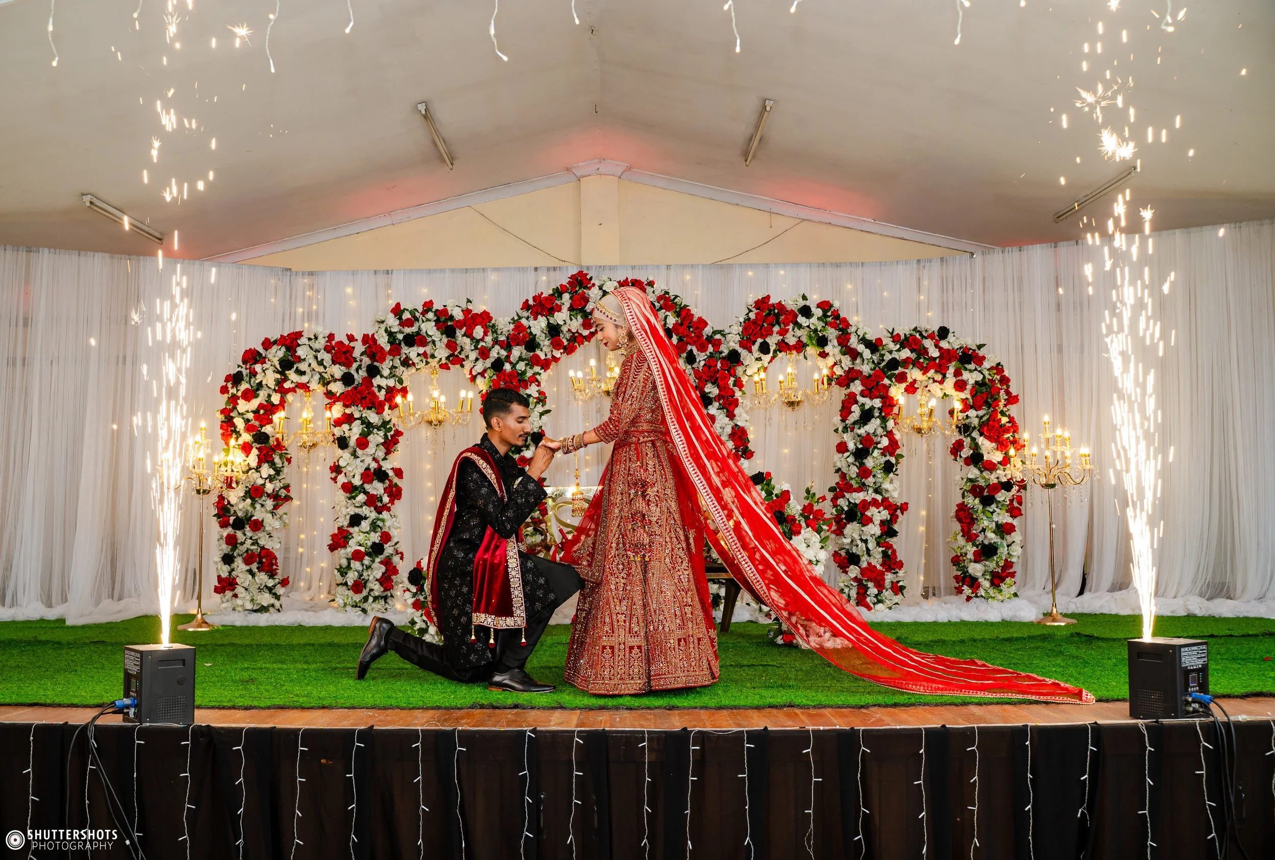 A couple getting engaged during a wedding celebration on a decorated stage with red and white flowers, candles, and sparklers.