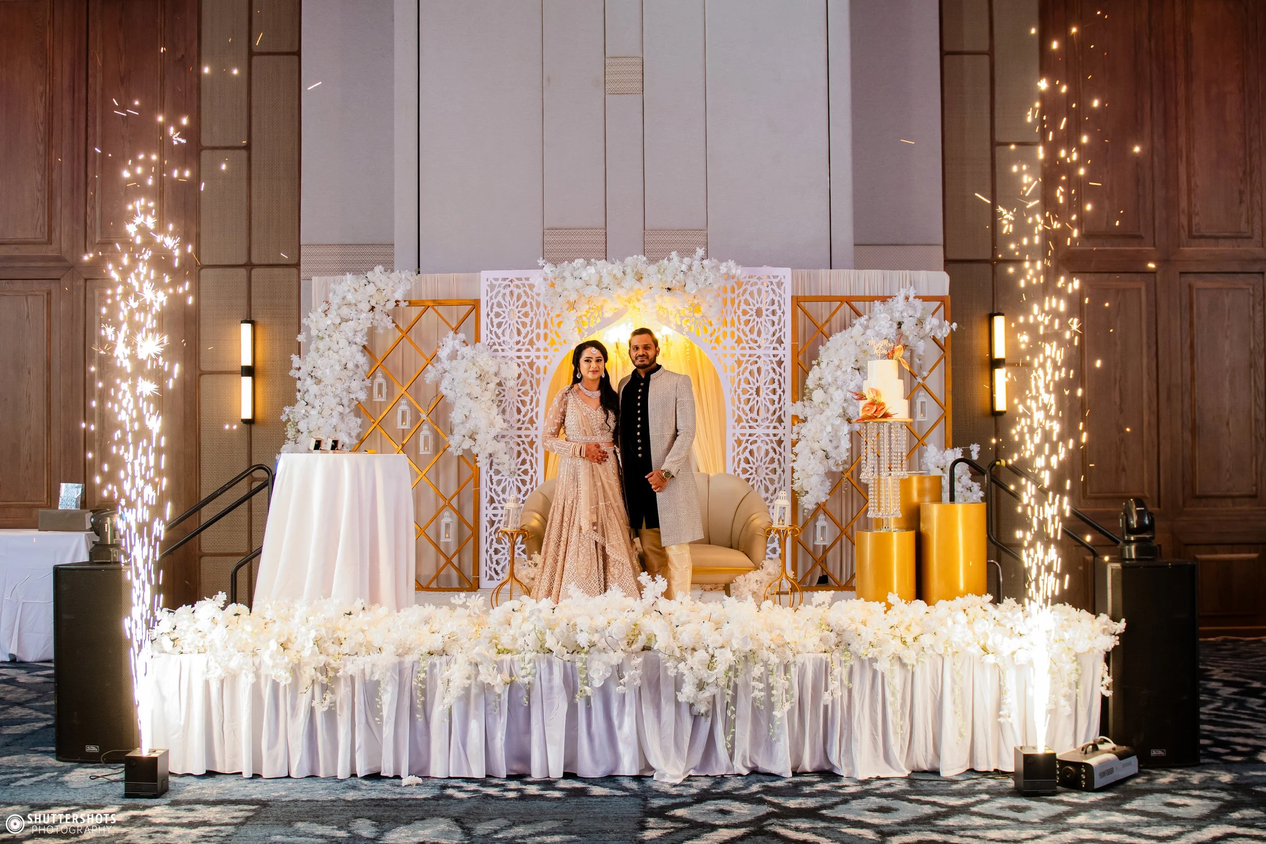 A couple in traditional Indian wedding attire standing on a decorated stage with white flowers, gold accents, and sparklers.