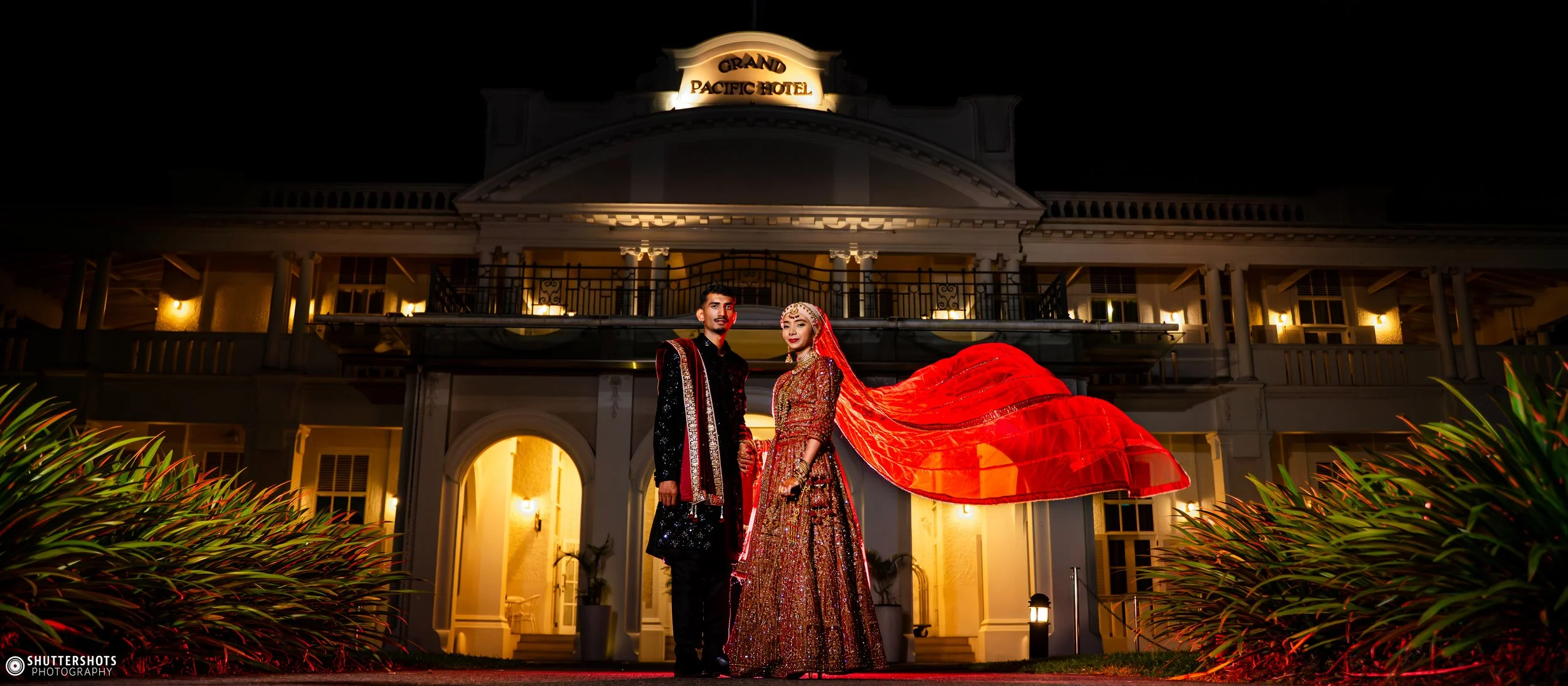 An Indian bride and groom in traditional attire standing in front of the Grand Pacific Hotel at night, with illuminated building and greenery around them.