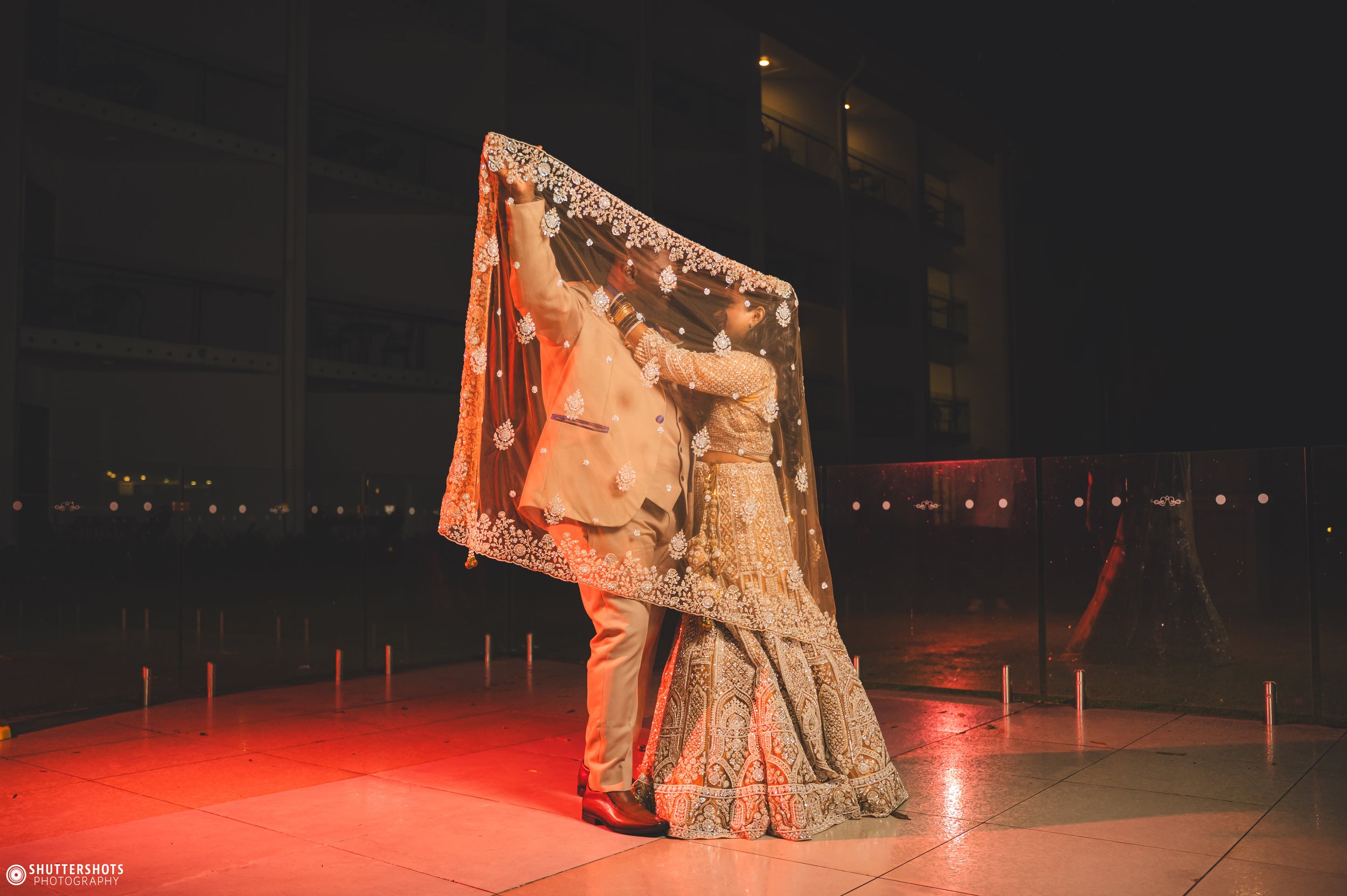 A newlywed couple is dancing at nighttime on a balcony with a glass railing. The groom is lifting the bride's veil, which is decorated with intricate embroidery. The bride is wearing a richly embroidered lehenga, and the groom is in a beige suit. The background shows a dark night sky and city lights reflection on the glass.