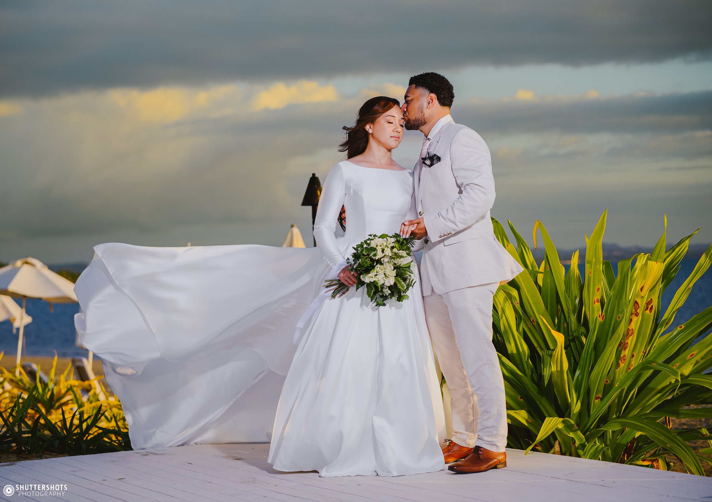 A bride and groom stand close together on a beach, with the ocean and cloudy sky in the background. The bride is holding a bouquet of white flowers, wearing a long white wedding dress, and the groom is wearing a light-colored suit. The groom is kissing the bride on her forehead, and they are holding hands.
