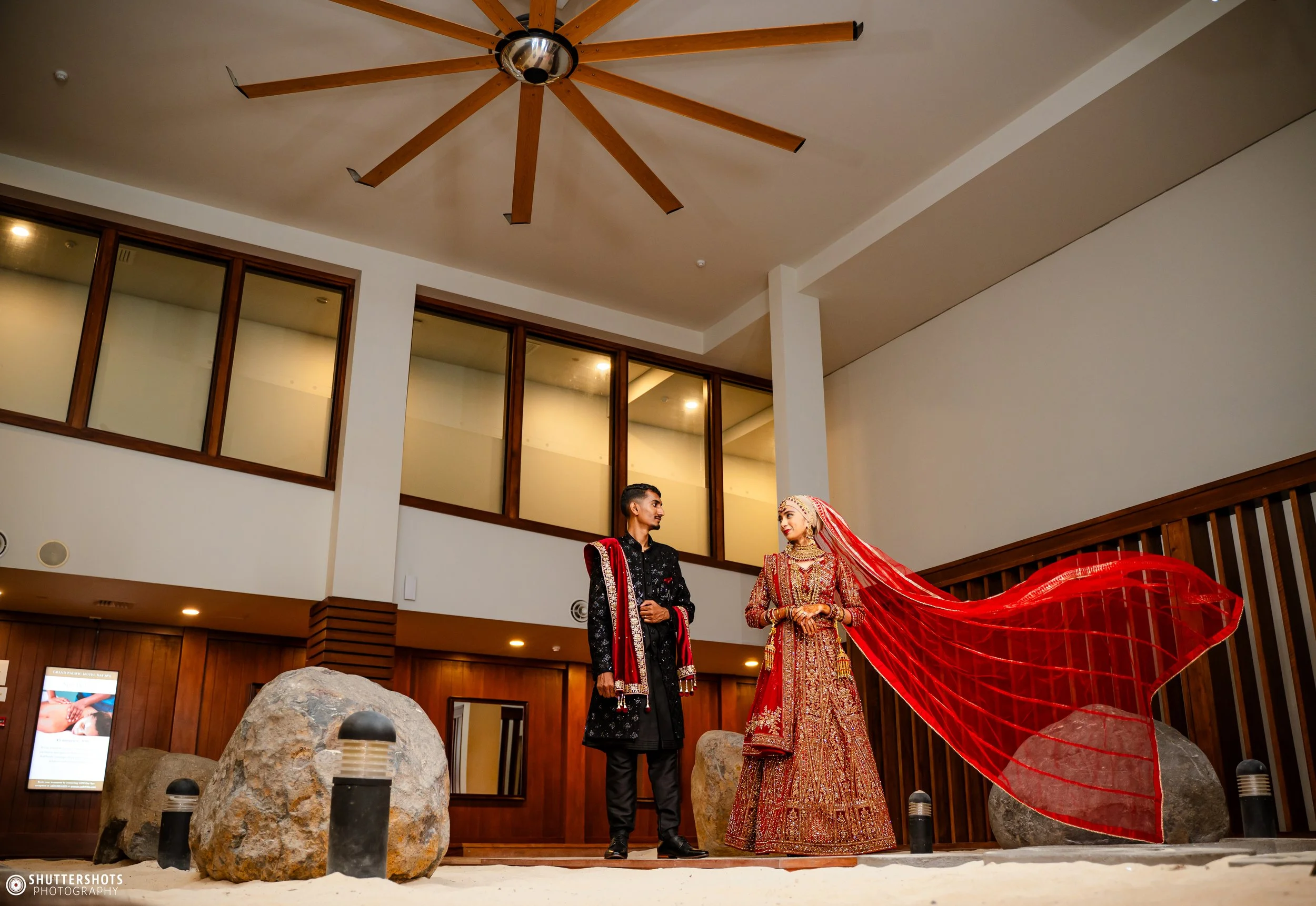 A bride and groom in traditional Indian wedding attire standing indoors in front of a wooden wall, with large rocks and small lights on the floor.