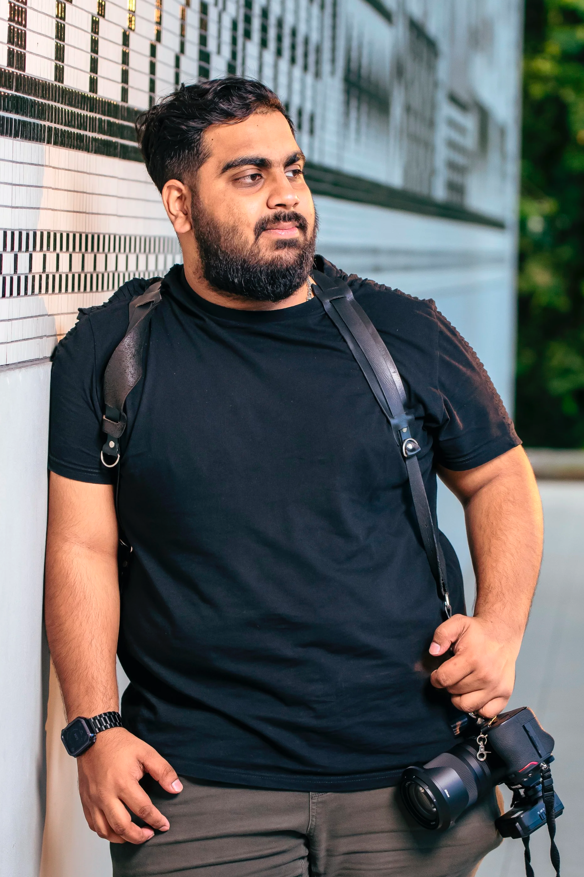 A man with a beard and mustache, wearing a black t-shirt and khaki pants, is leaning against a decorative tiled wall. He is carrying a black camera with a strap around his neck and is wearing a black smartwatch on his left wrist.