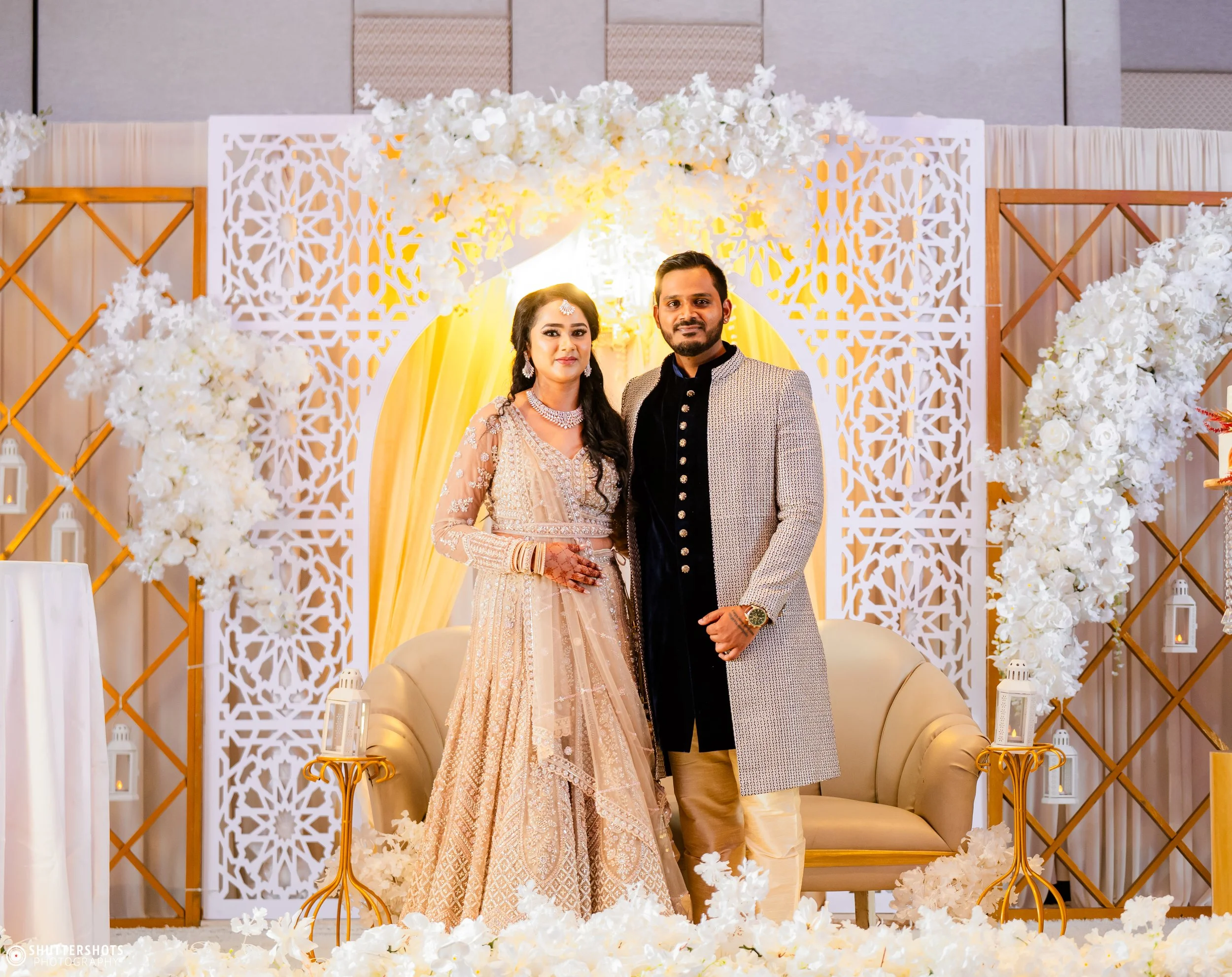 A newlywed couple dressed in traditional Indian wedding attire standing in front of a decorated wedding stage with white flowers, yellow drapery, and ornate white lattice panels, posing for a photo.