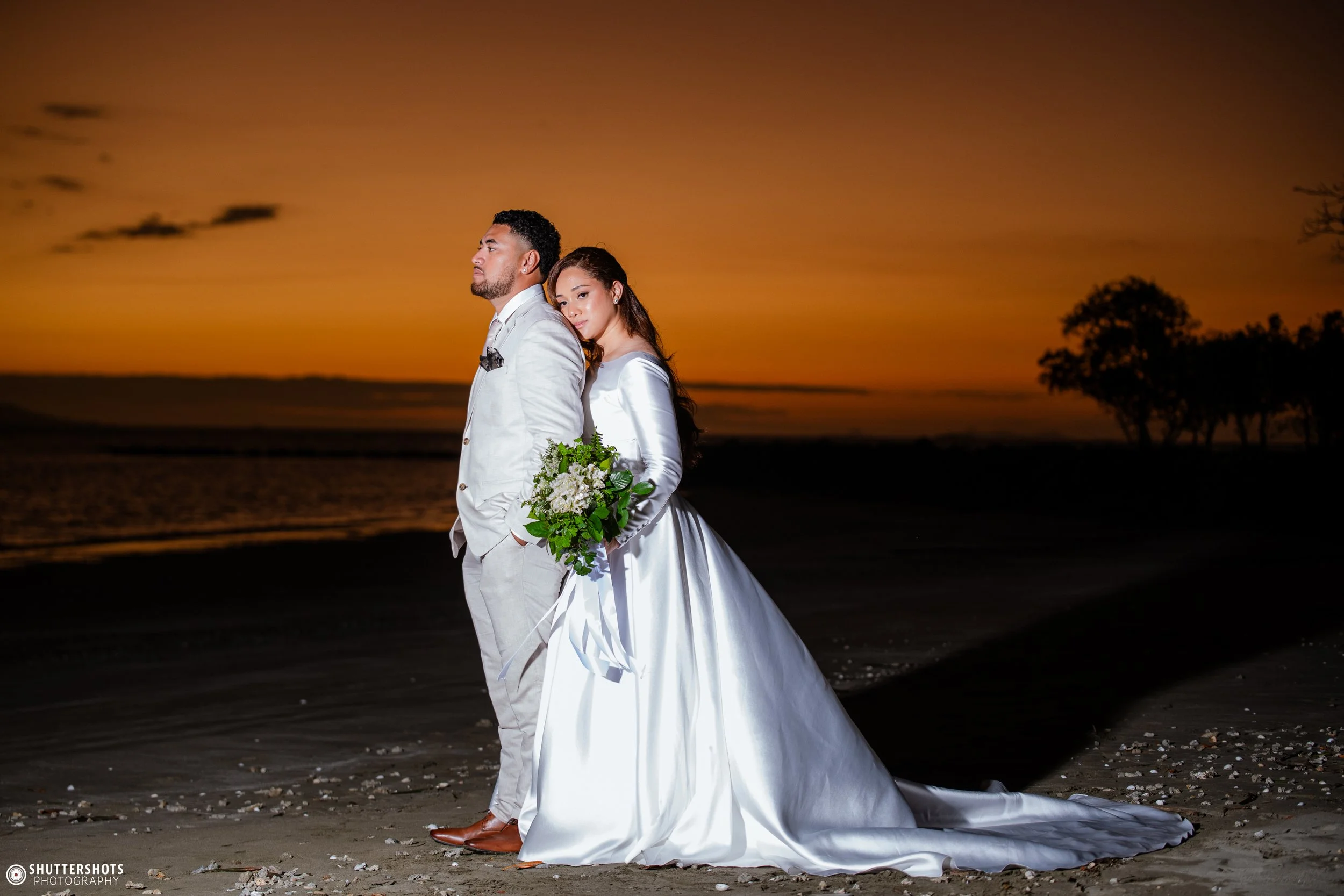 A bride and groom standing on a beach during sunset, with the bride holding a bouquet of flowers and the groom with his hands in his pockets, both dressed in white wedding attire.