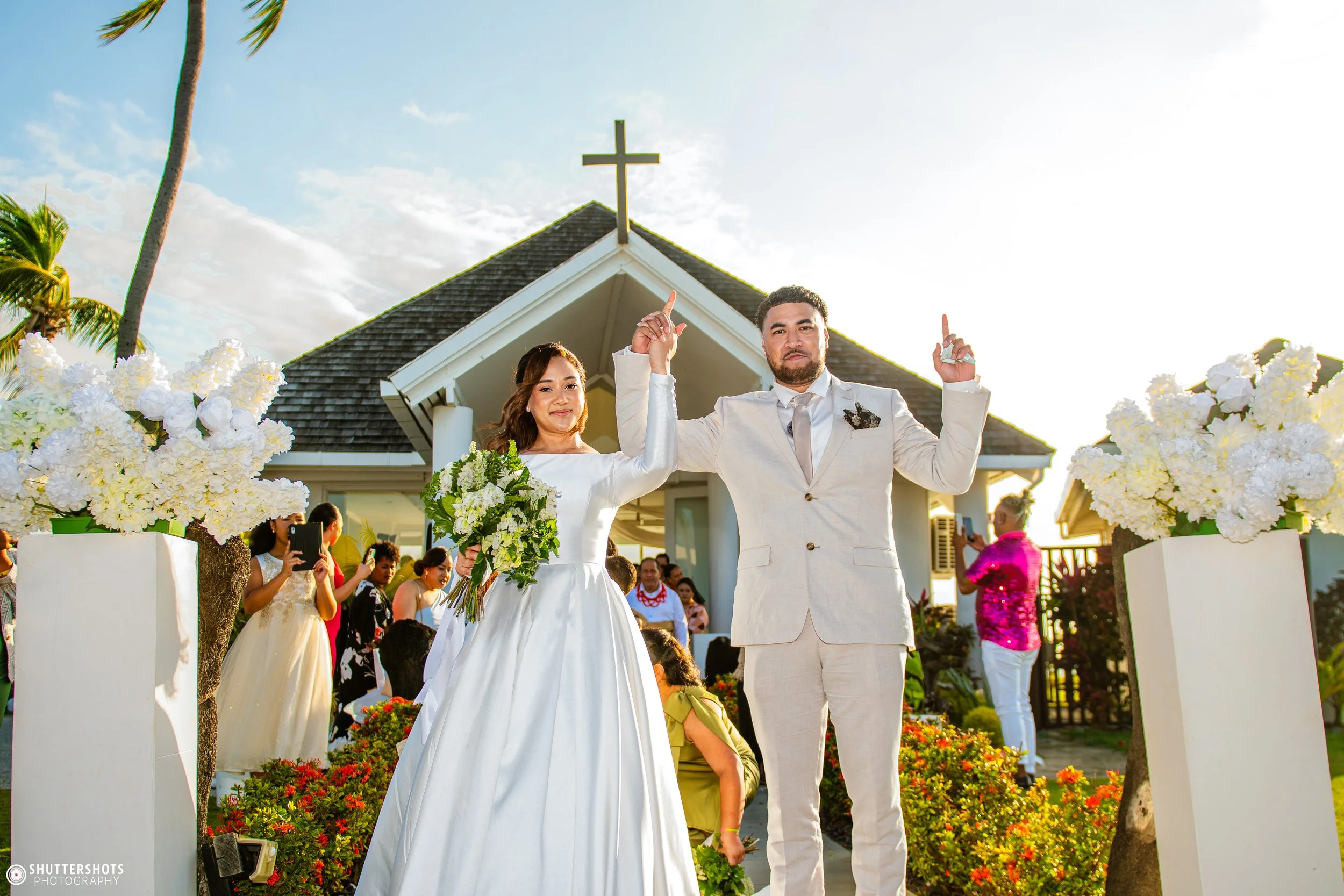 A couple dressed in wedding attire celebrating outside a church with white flowers and guests in the background.
