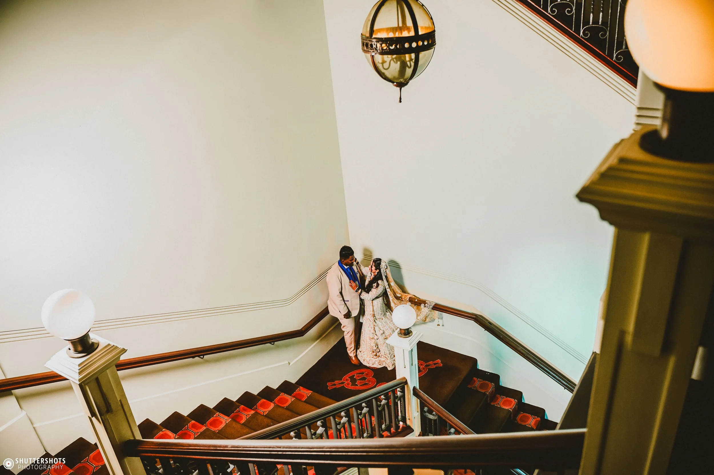 A couple dressed in wedding attire standing at the top of a staircase, looking at each other, with blank white wall behind them and decorative lighting fixtures above.