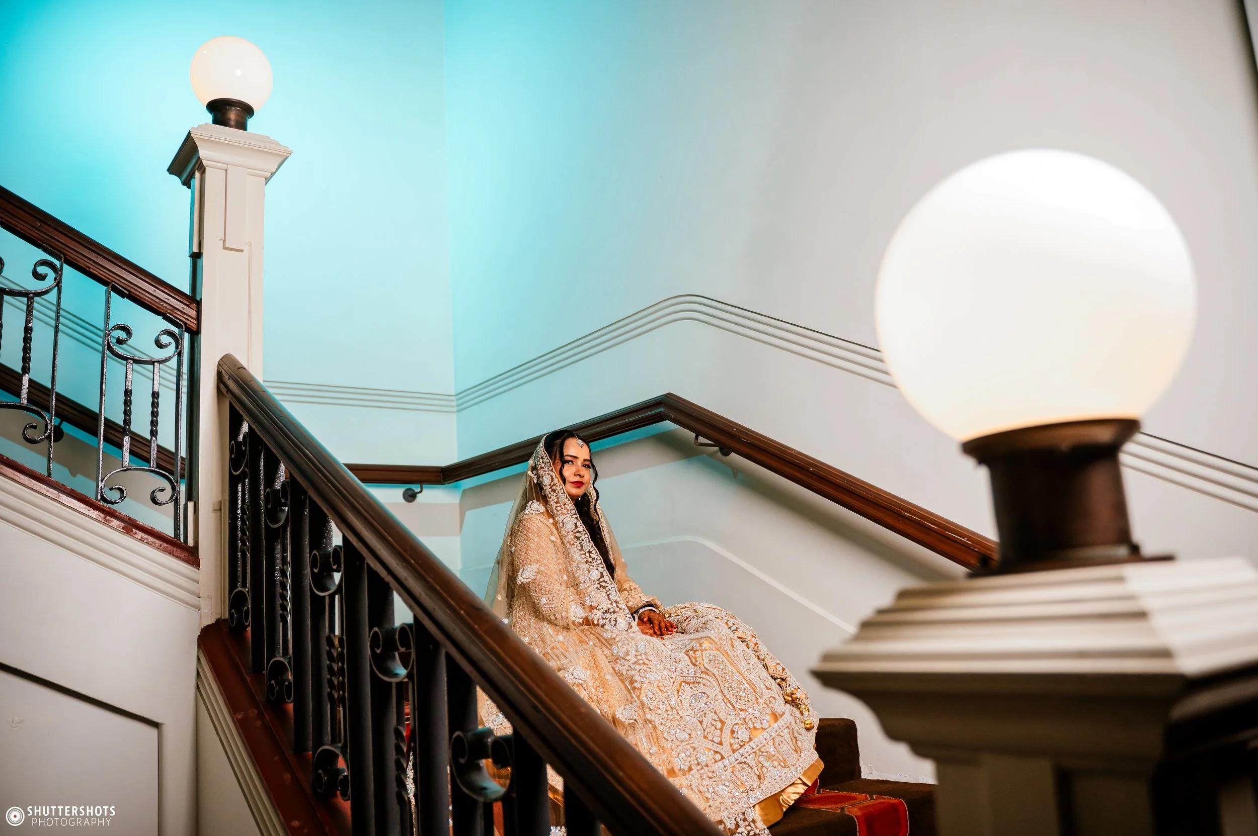 A woman in traditional Indian bridal attire sitting on a staircase inside a building with ornate railing and wall lamps.