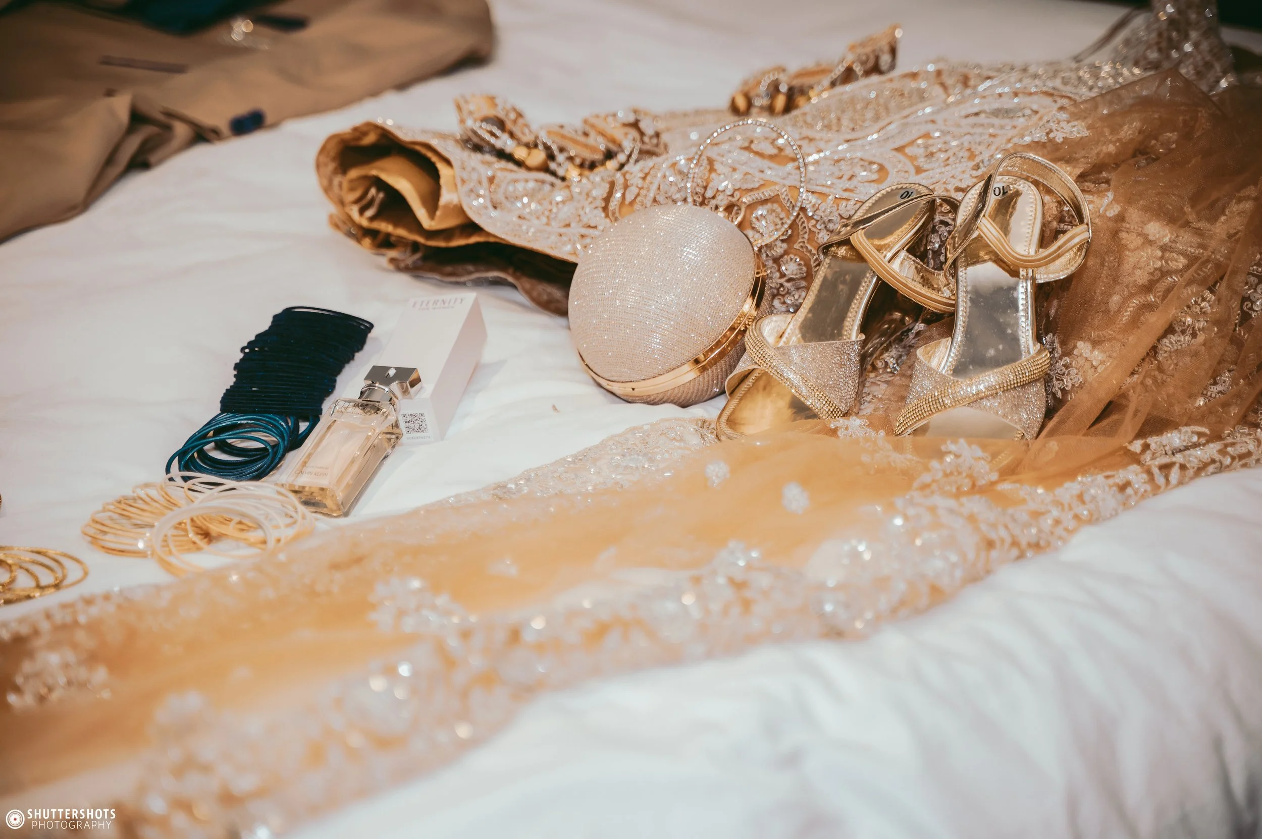 Elegant wedding accessories on a white bed, including a pair of gold and silver high heel shoes, a round sparkling clutch purse, gold jewelry, a perfume bottle, and a decorative lace fabric.
