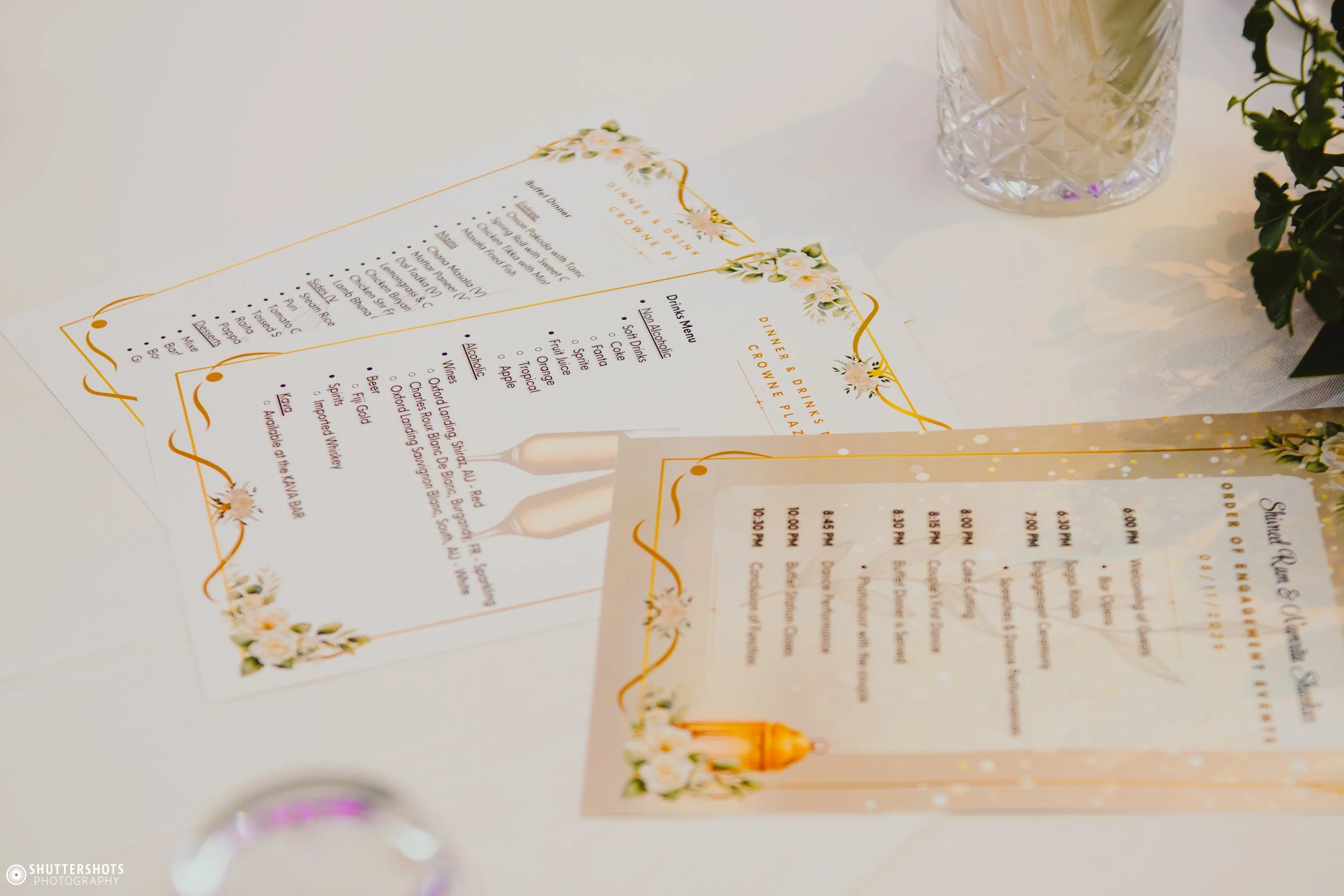Two printed menus on a table for a dinner and drinks event, decorated with floral borders, a glass of ice with lemon slices, and a small potted plant nearby.