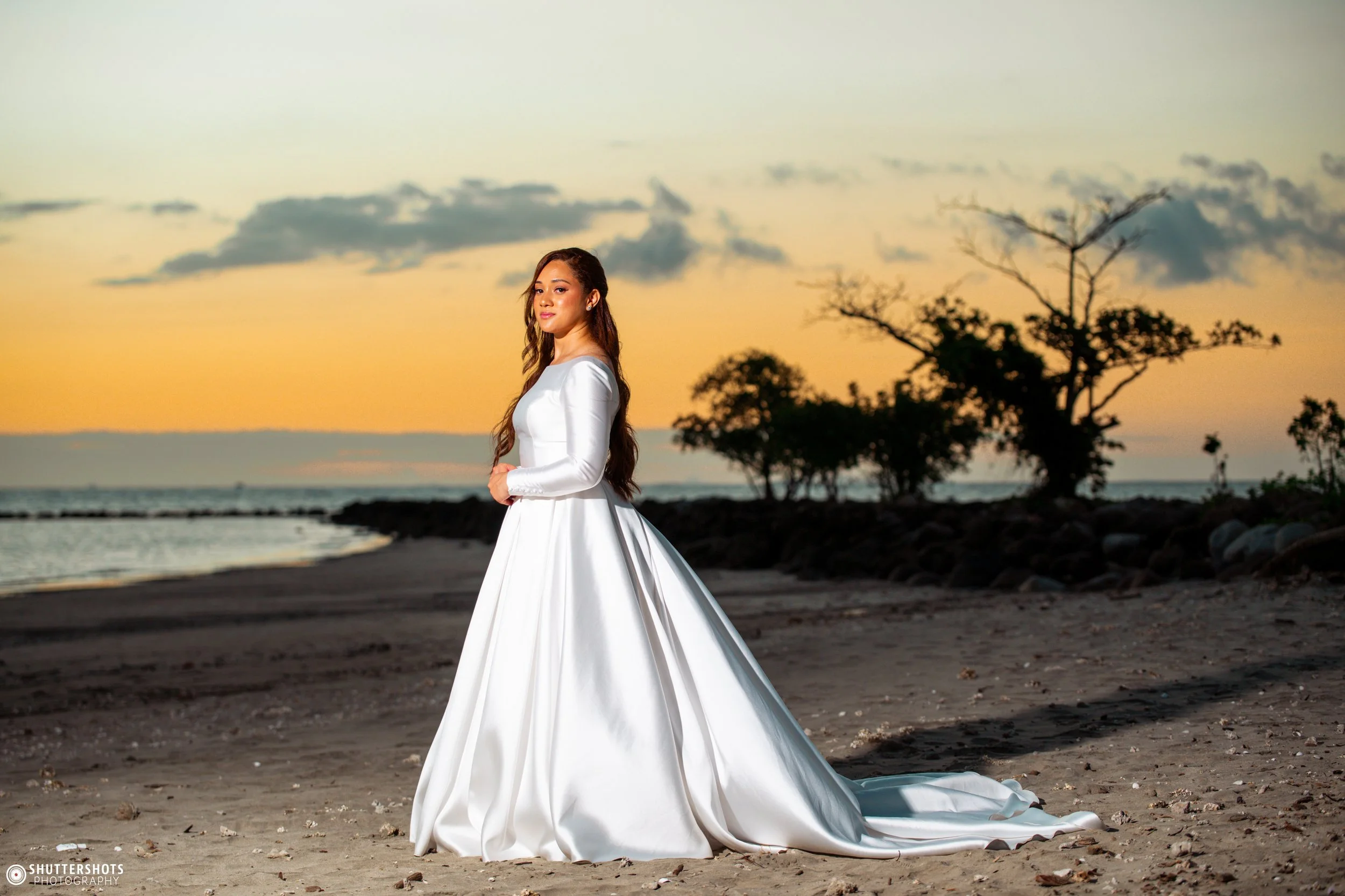 A woman in a white wedding dress standing on a beach at sunset