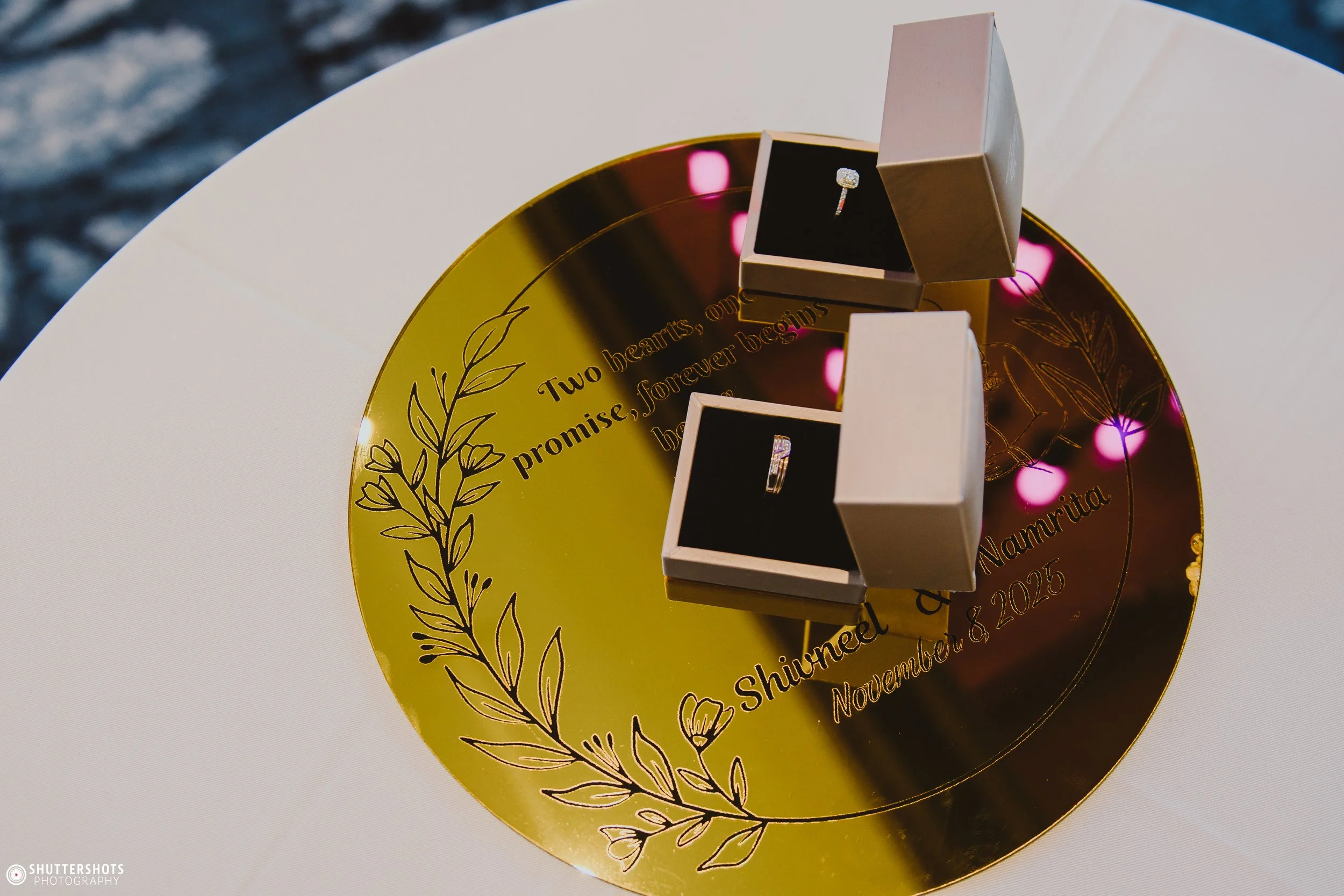 Two wedding rings in open boxes placed on a gold circular plaque with engraved text and decorative floral designs.