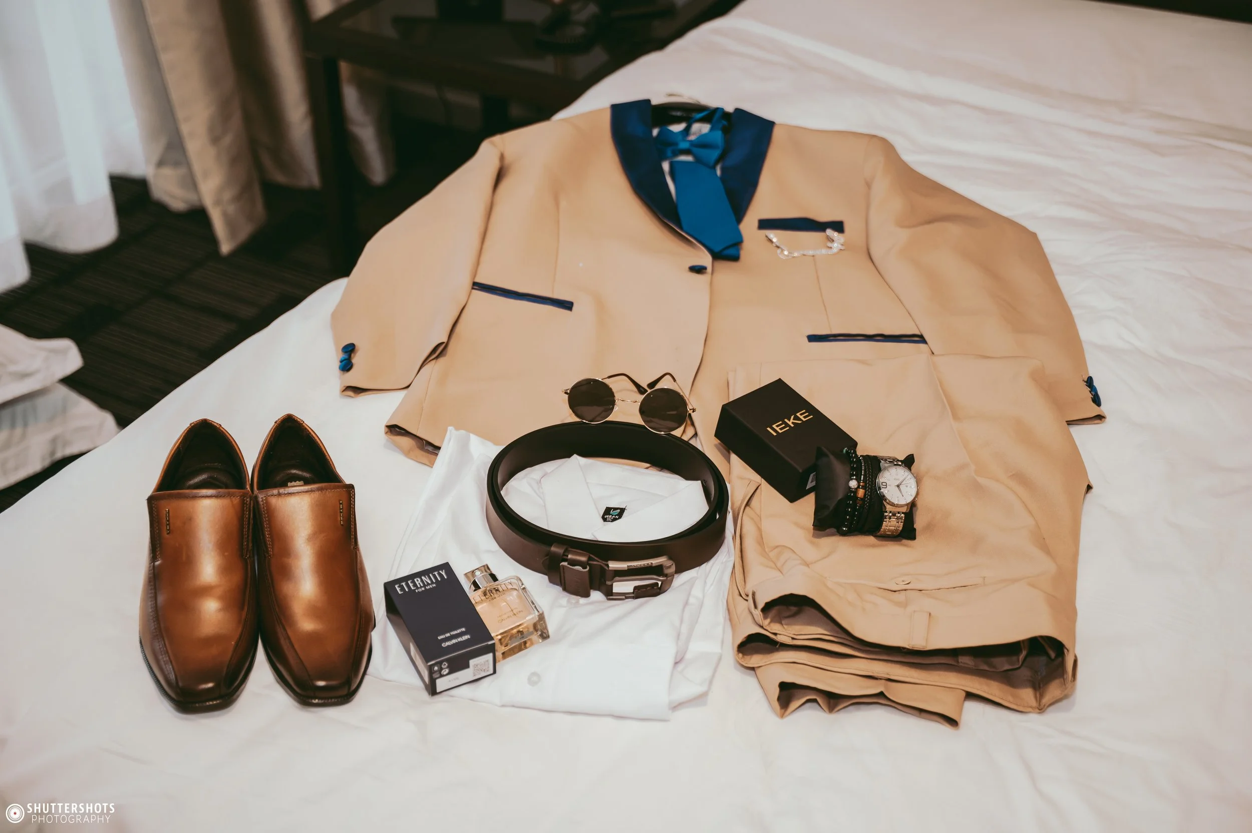 A beige suit jacket with blue trim and a blue necktie, beige trousers, a pair of brown leather shoes, round sunglasses, a belt, a watch, a pack of cigarettes, a perfume bottle, and a box, all arranged on a white sheet on a bed.