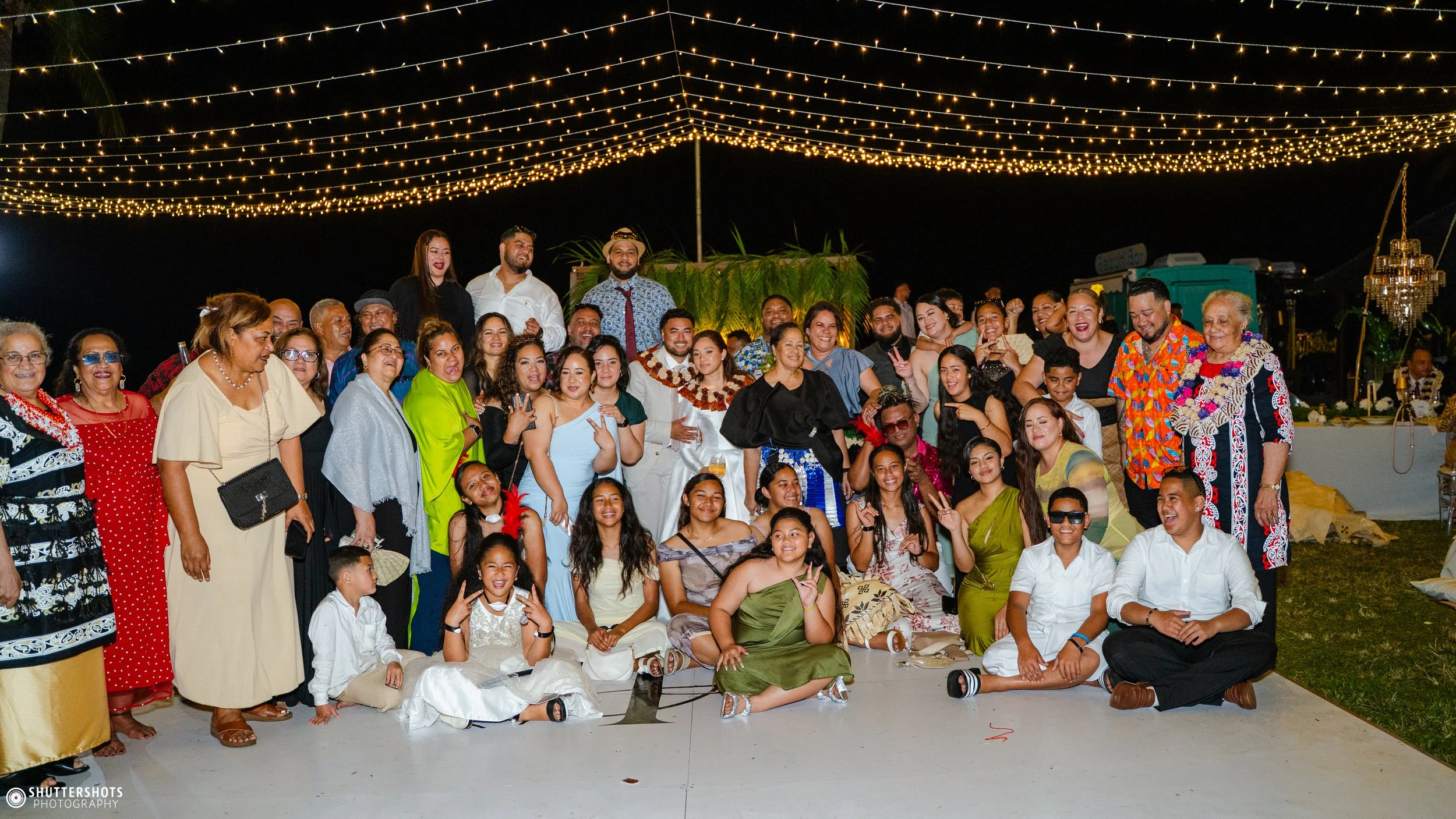 Large group of people at night under string lights, celebrating at a festive outdoor event, some wearing traditional and colorful attire, smiling, and posing for a group photo.