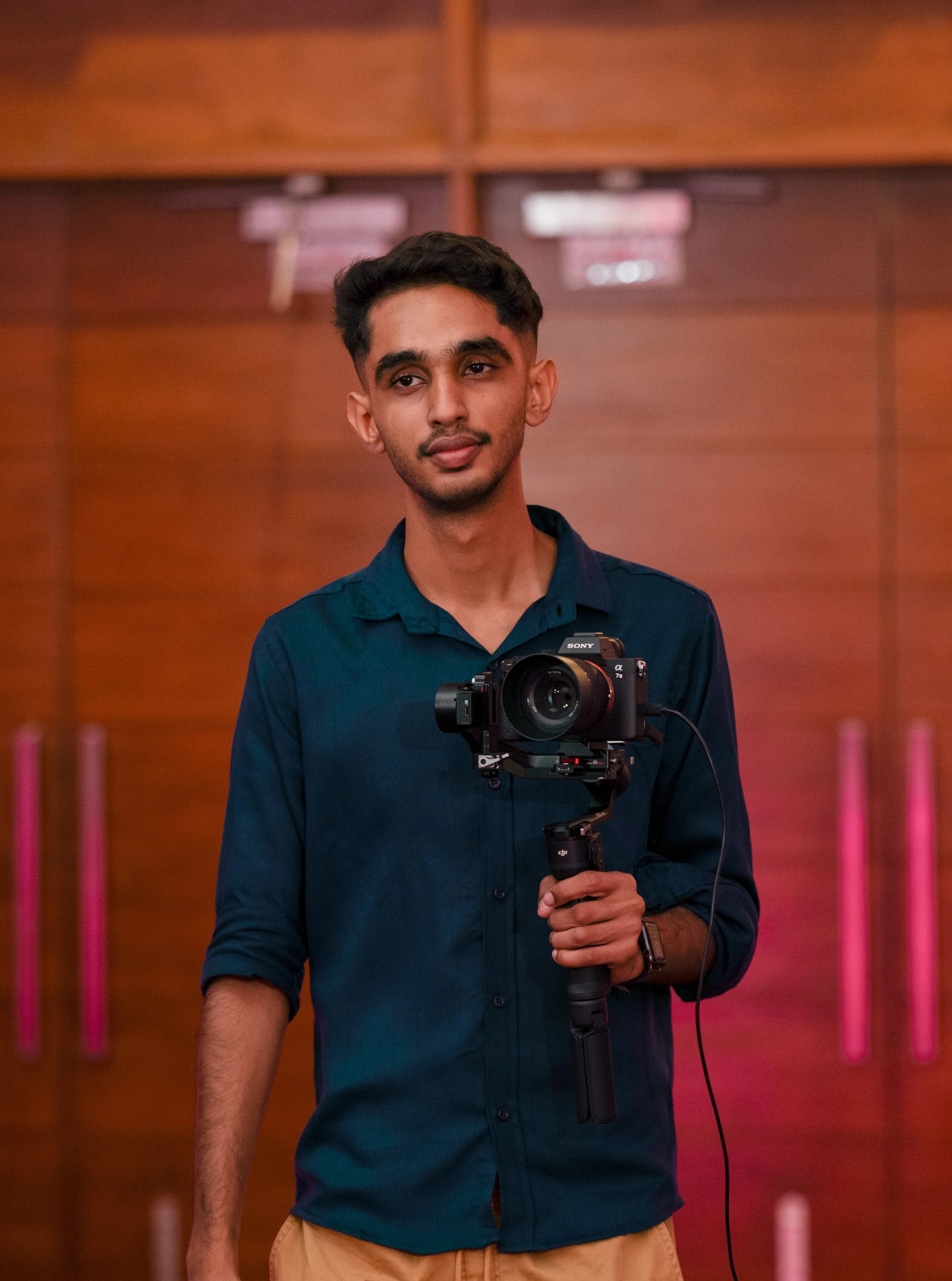 A young man holding a camera on a gimbal, standing indoors with a wooden wall background.