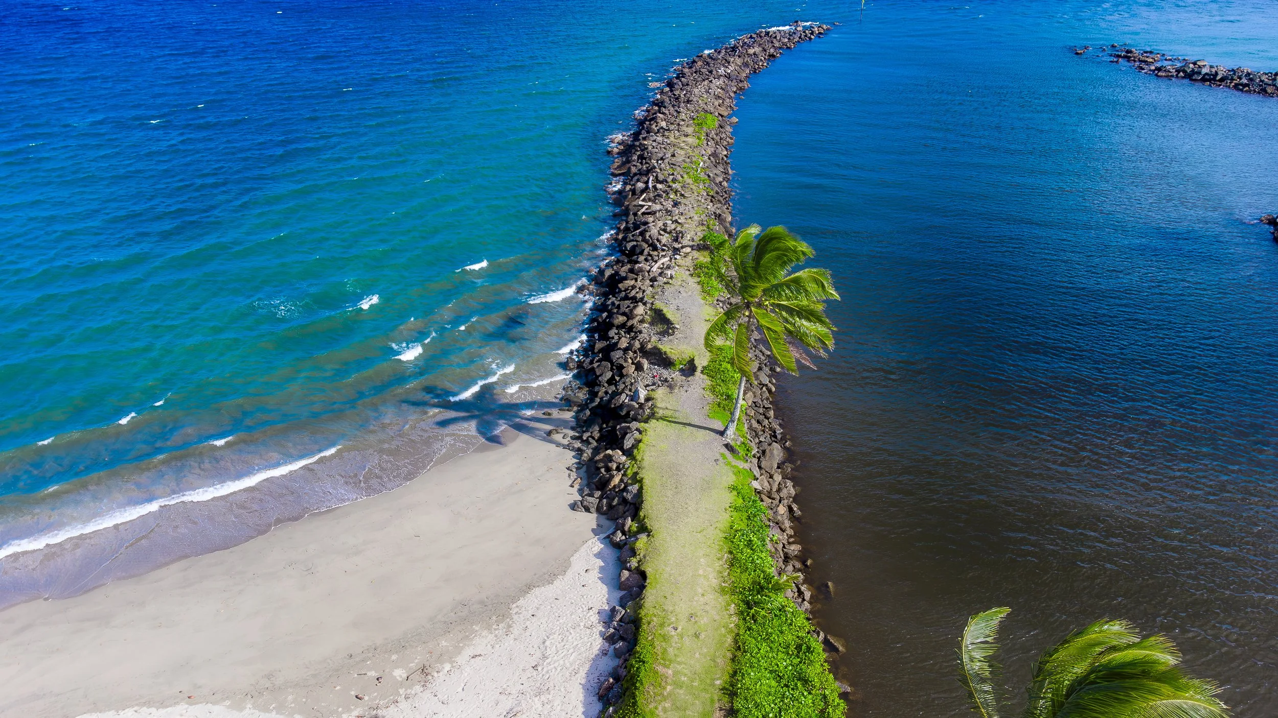 Aerial view of a split island with one side having white sandy beach and turquoise water, and the other side dark blue water, divided by a narrow grassy path with palm trees.