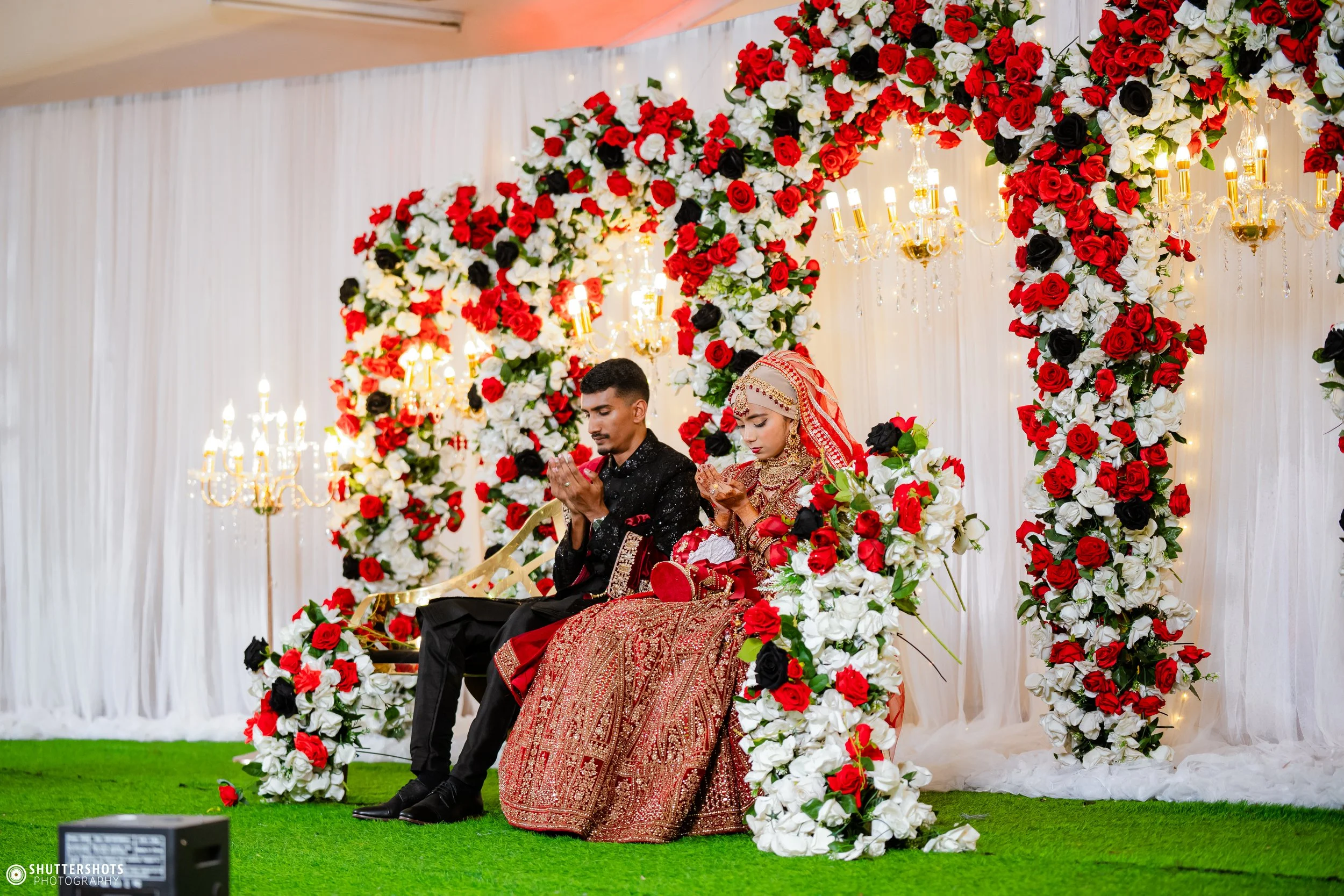 A bride and groom sitting on a throne-like chair at their wedding ceremony, surrounded by a floral arch with red, white, and black flowers, and lit chandeliers in the background.