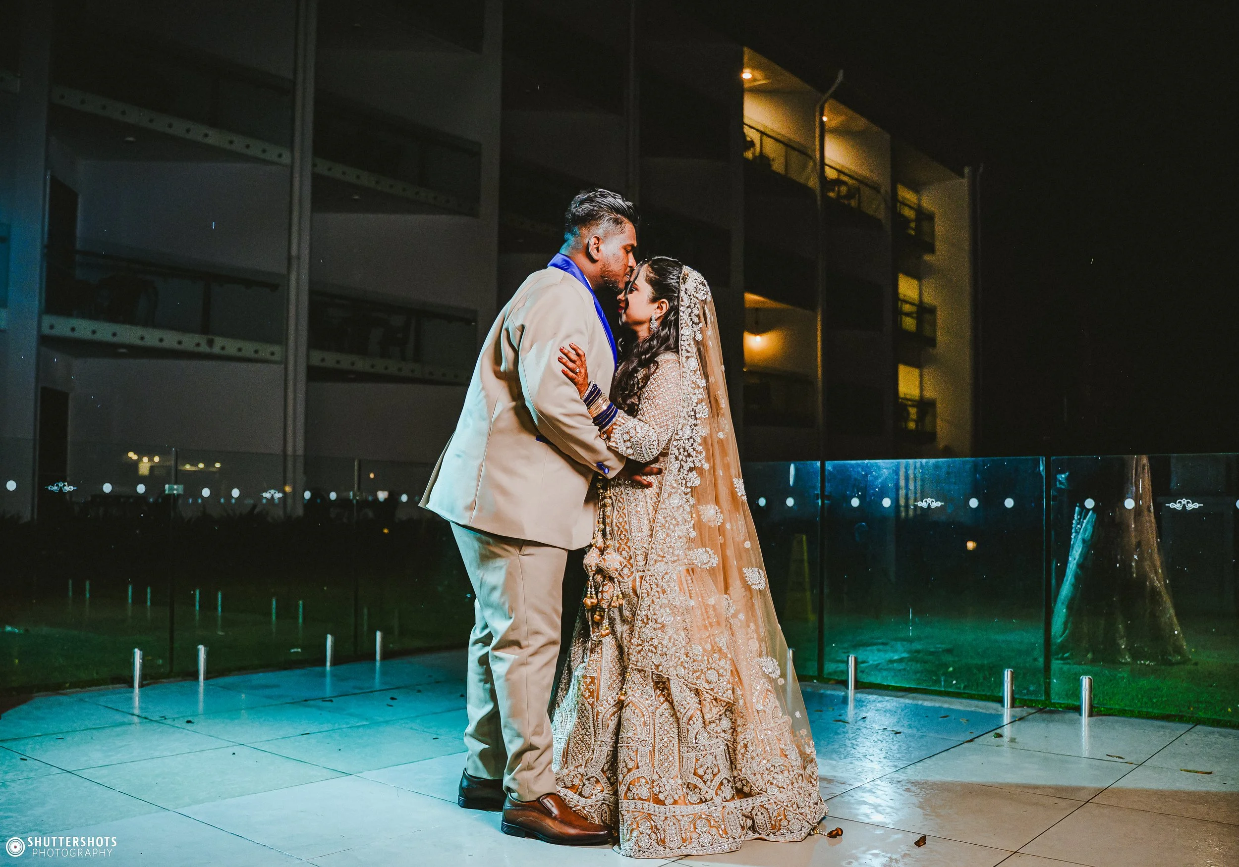 A couple dressed in traditional wedding attire sharing a close moment on a balcony at night, with a modern building in the background.