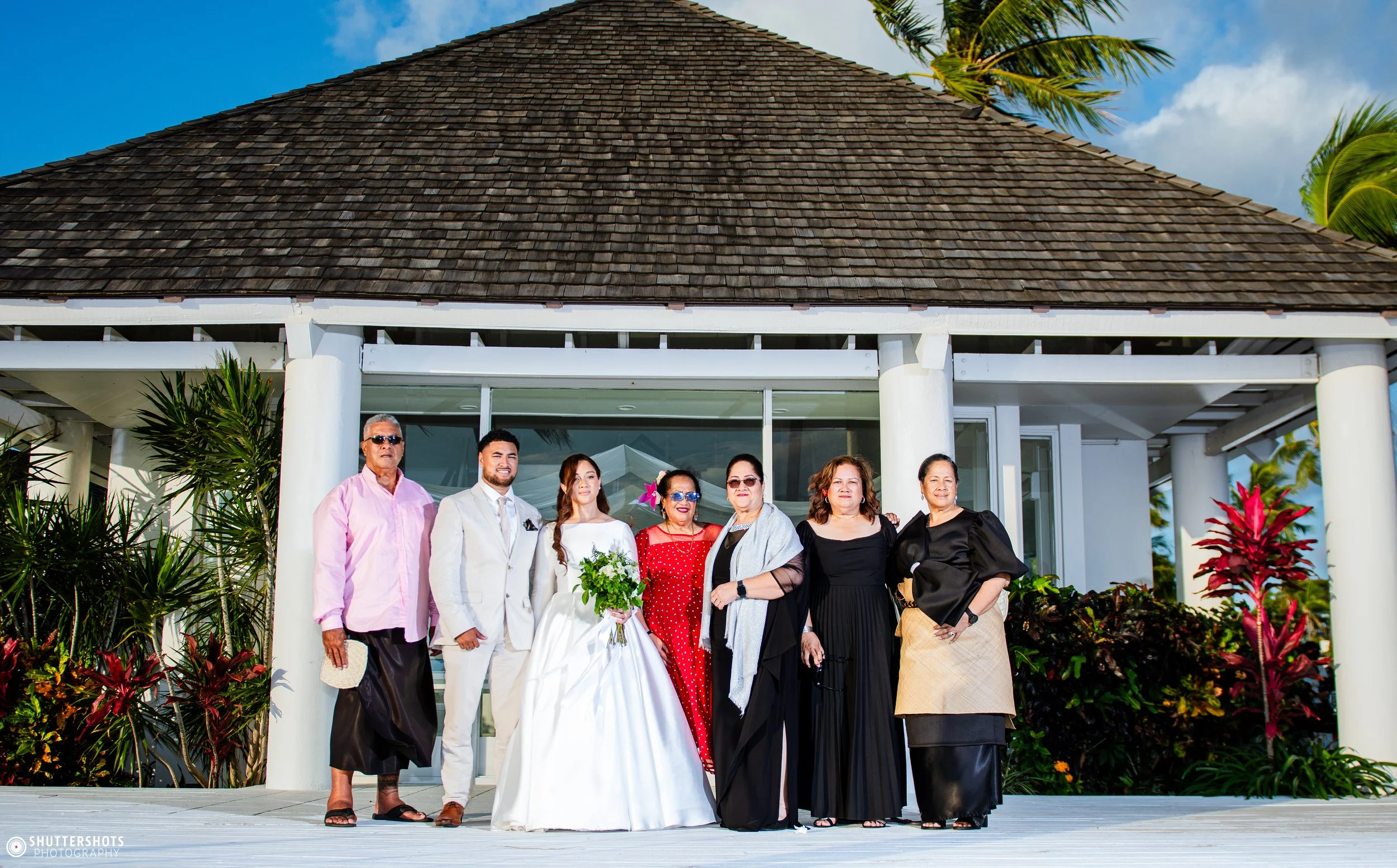 A group of seven people standing in front of a modern house with tropical plants, during daytime. One woman in a white wedding dress holding a bouquet, indicating a wedding celebration.
