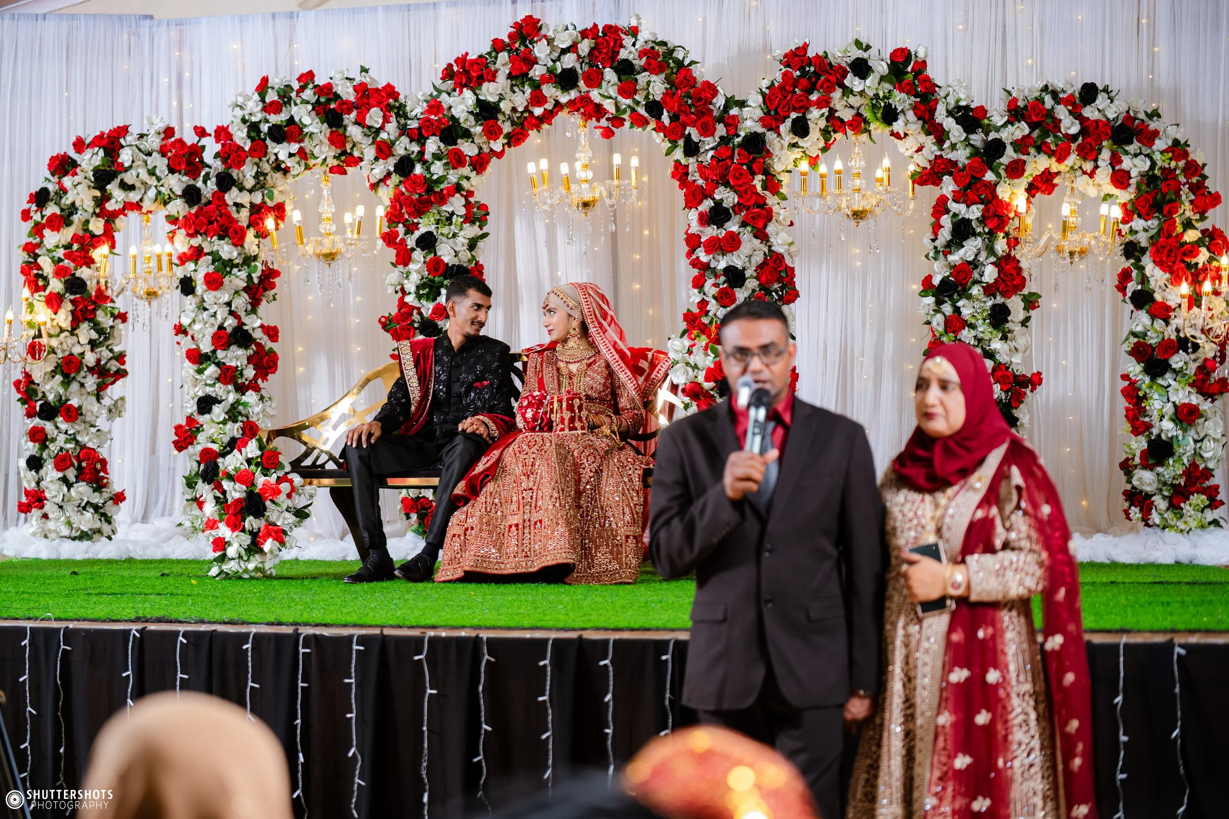A bride and groom sitting on a swing decorated with red and white flowers during a wedding ceremony, with two people speaking at microphones in the foreground.