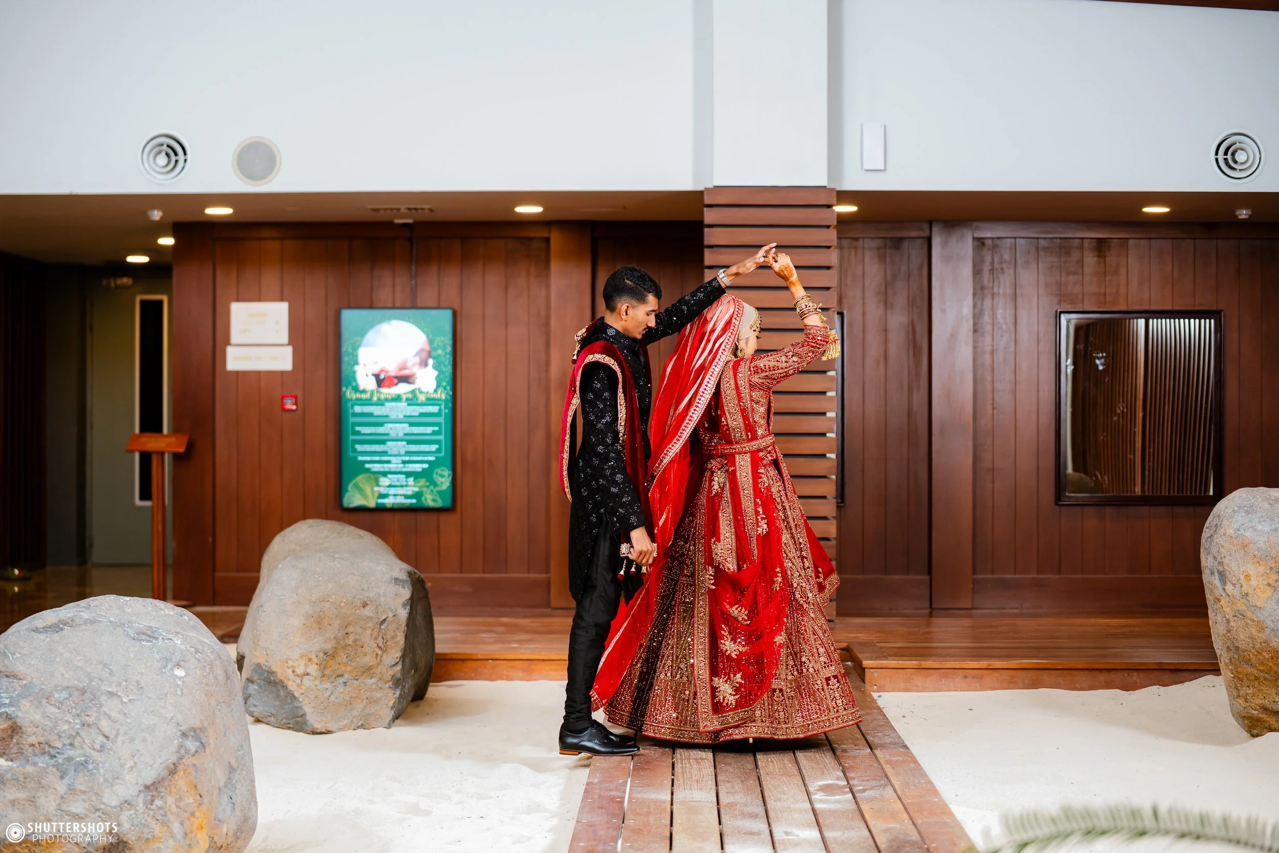 A newlywed couple dressed in traditional Indian wedding attire dancing indoors, with the groom spinning the bride.