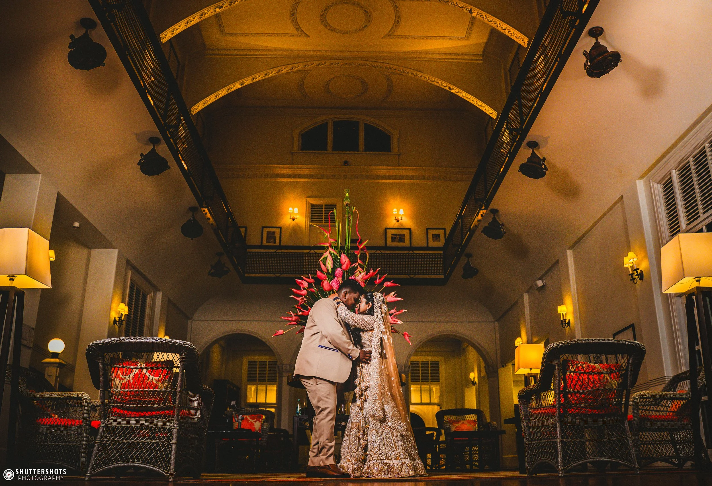 A bride and groom embrace in a warmly lit indoor space with elegant decor, dark wicker chairs with red cushions, and a large floral arrangement behind them.