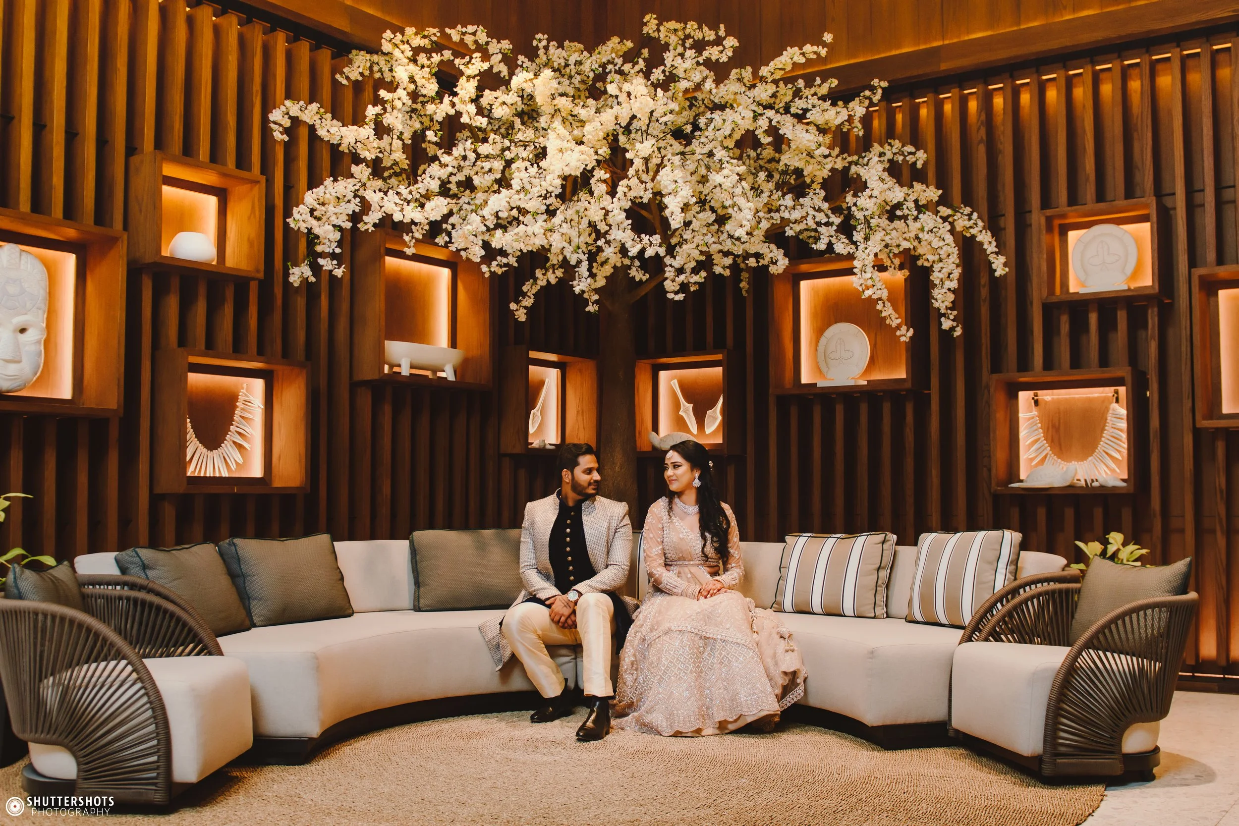 A man and woman dressed in traditional Indian attire sitting on a modern white sectional sofa in a decorated interior with a large floral tree and artwork displayed in illuminated shelves behind them.
