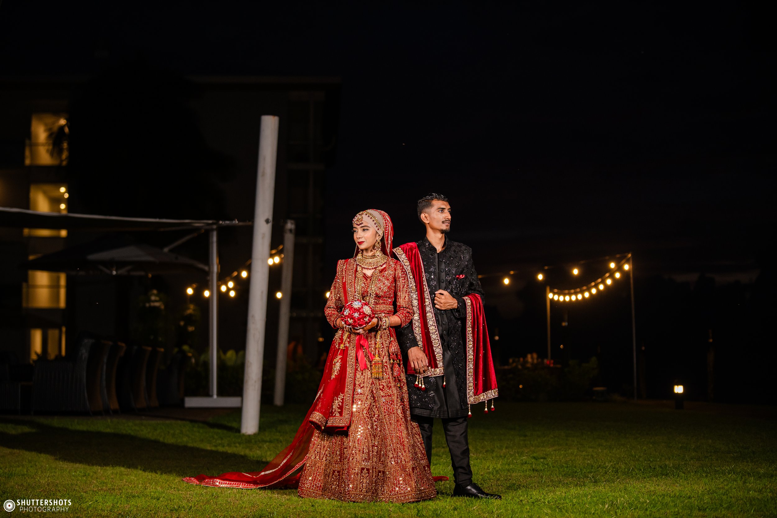 A bride and groom in traditional dark red and black wedding attire standing outdoors at night, with string lights in the background.