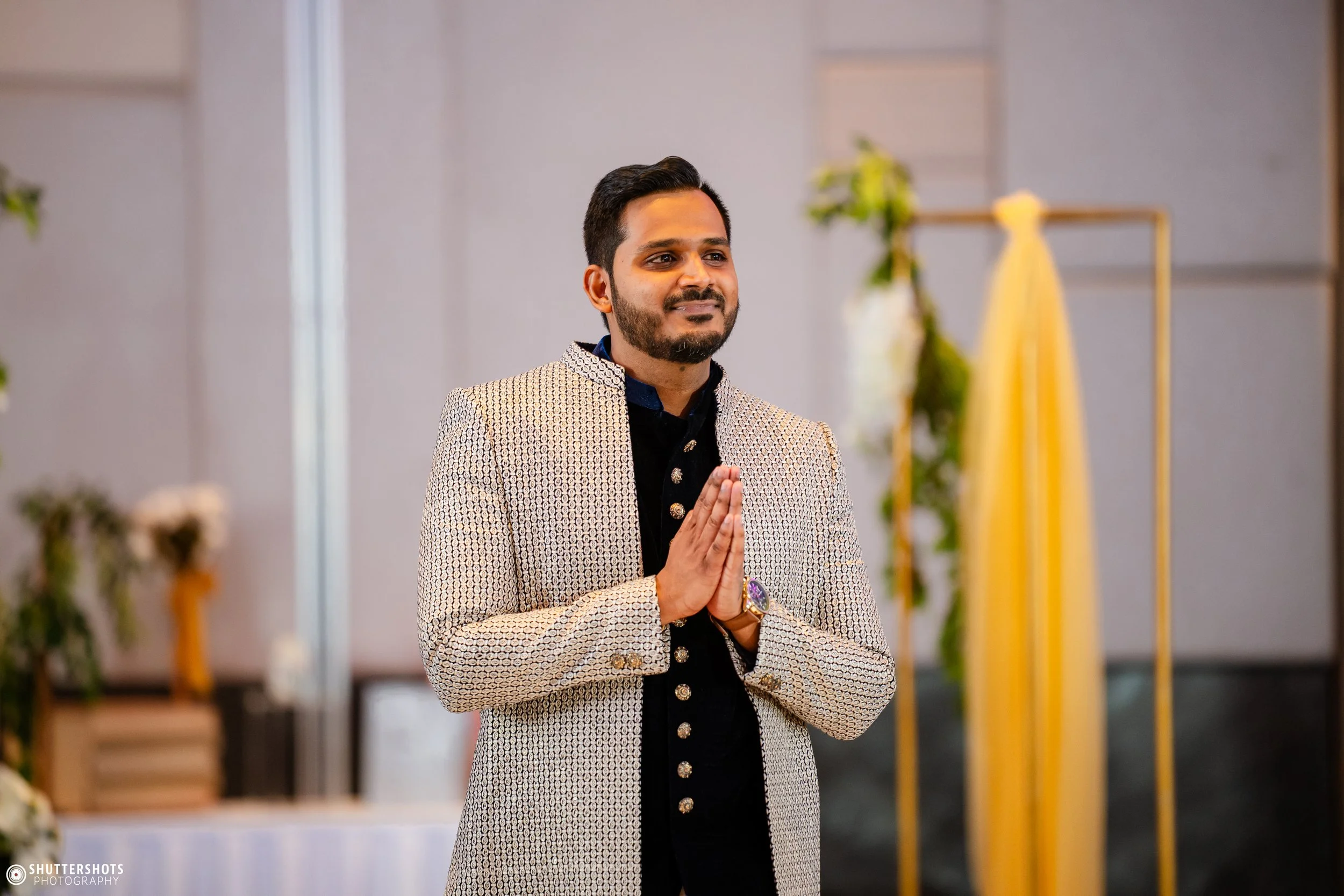 A man in traditional Indian attire standing with hands folded in prayer at a wedding or cultural ceremony.