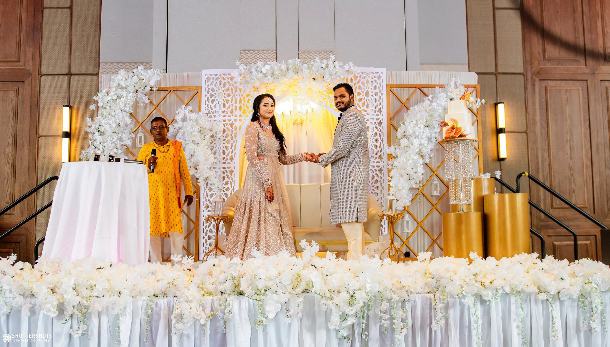 A couple in traditional wedding attire holding hands on a decorated stage with floral arrangements, a white loveseat, an officiant, and a tiered wedding cake.