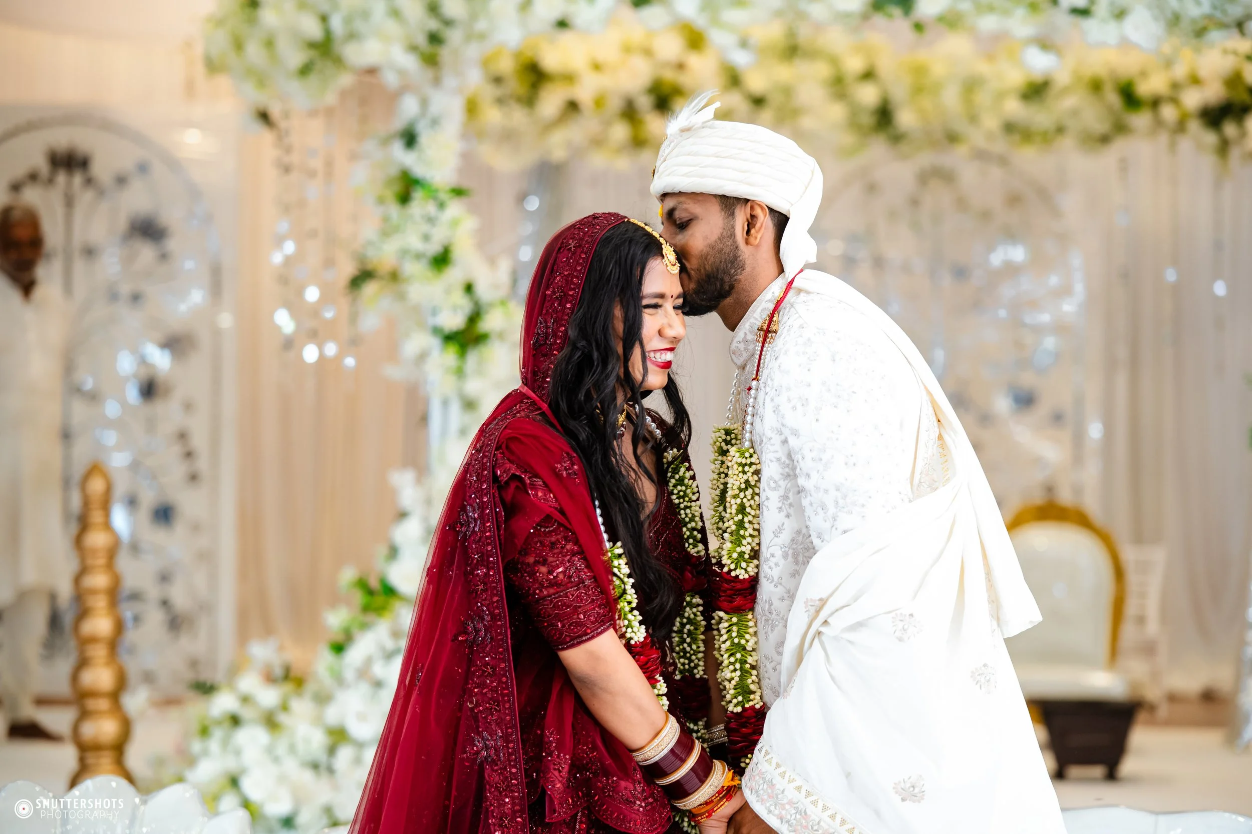 A bride and groom at their wedding ceremony, touching foreheads and smiling, with floral decorations and a white backdrop in the background.