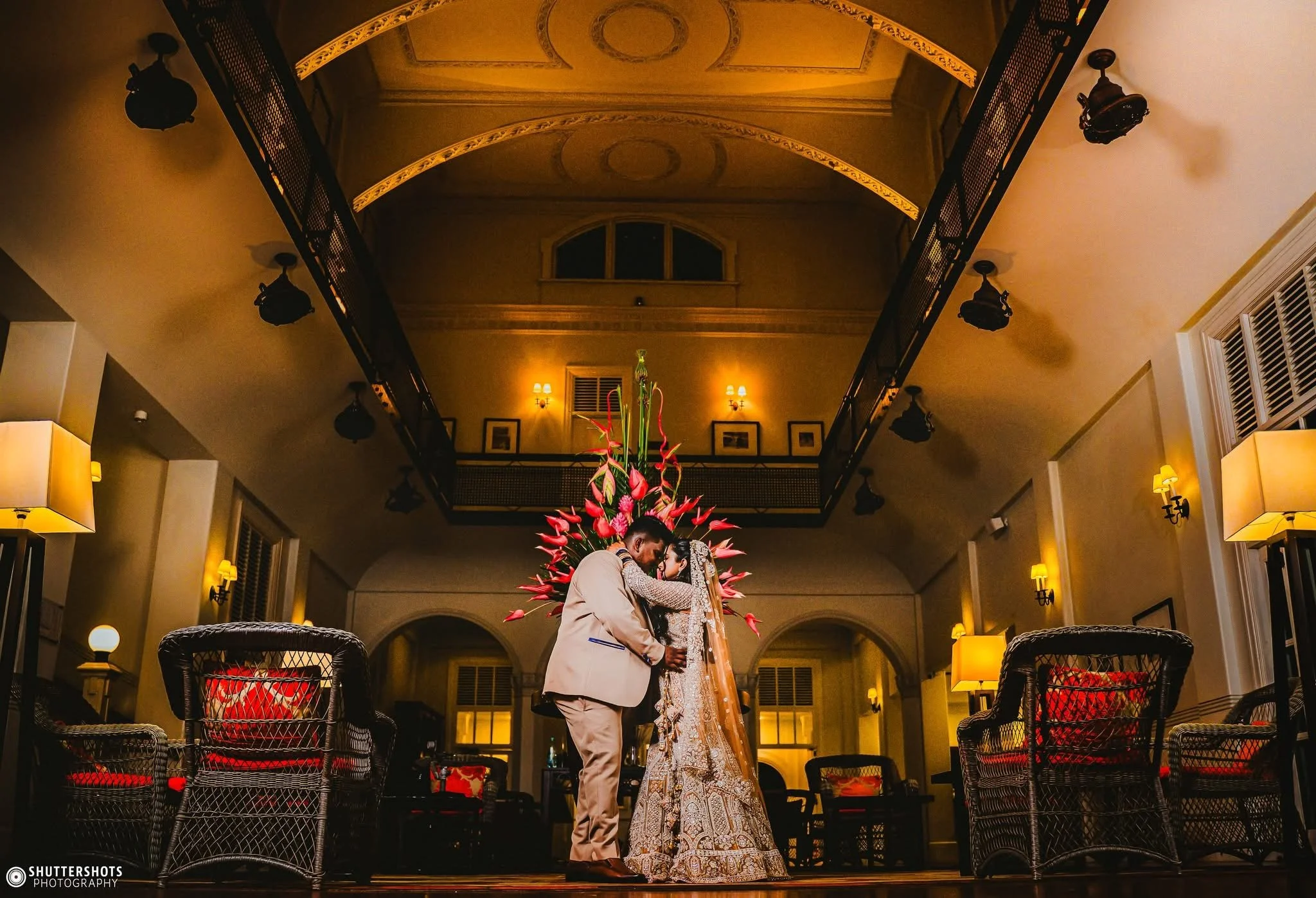 A couple dressed in traditional wedding attire sharing an intimate moment in a warmly lit, elegant indoor setting with a flower arrangement backdrop.