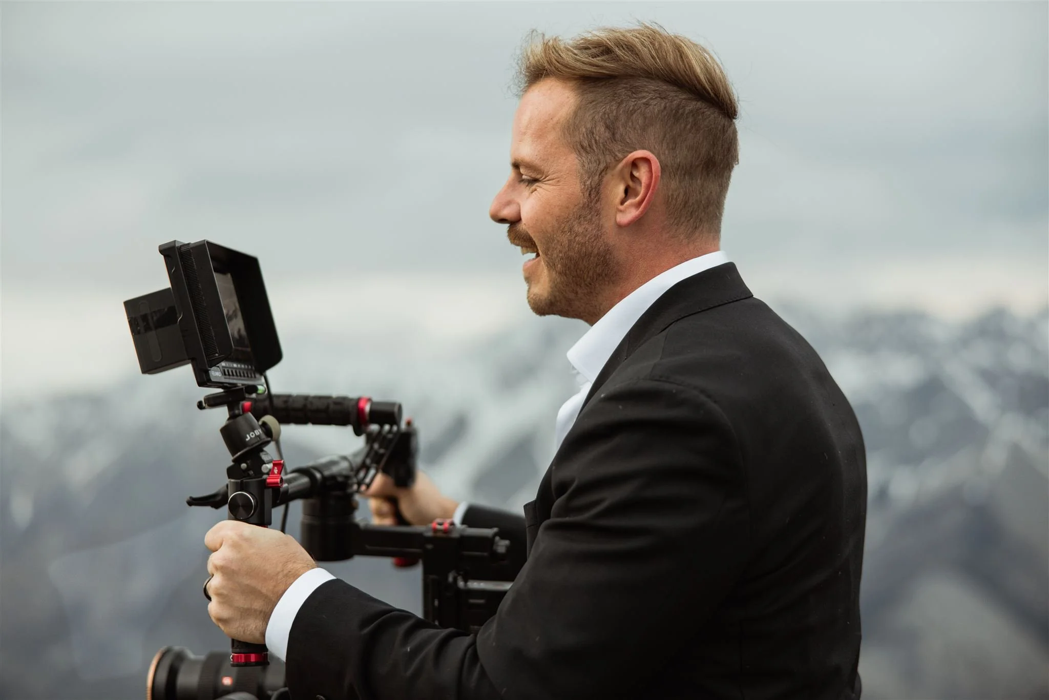 A man in a black suit holding a professional camera stabilizer with a mounted camera, outdoor mountain landscape in the background.